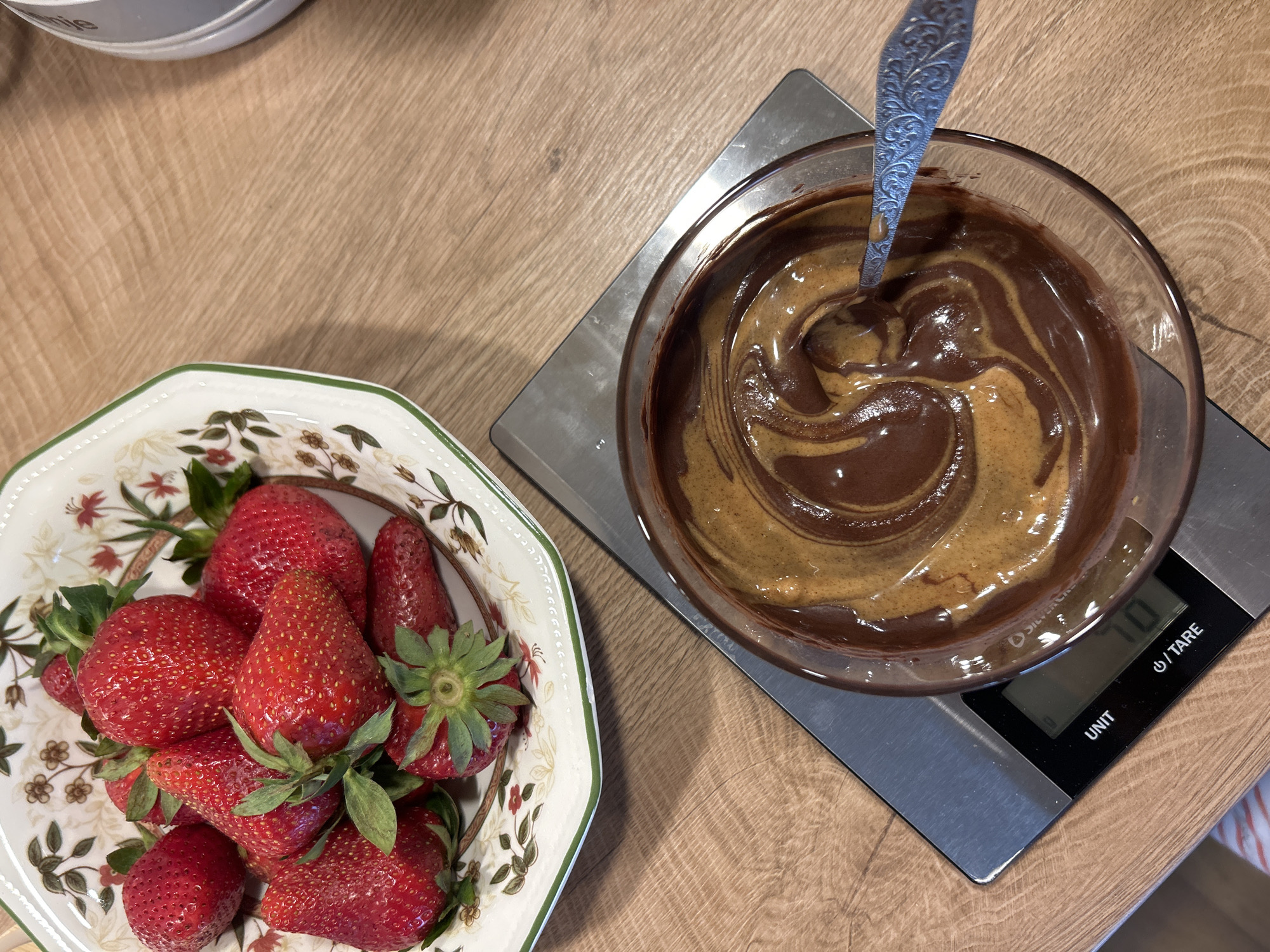 On a wooden surface, a bowl of strawberries sits to the left of a bowl of dark and light brown swirled liquid atop a digital scale. The scale reads "70". A silver spoon rests inside the liquid-filled bowl. The scene is likely indoors, but location cannot be confirmed as Plovdiv, Bulgaria without further information. No people are visible.