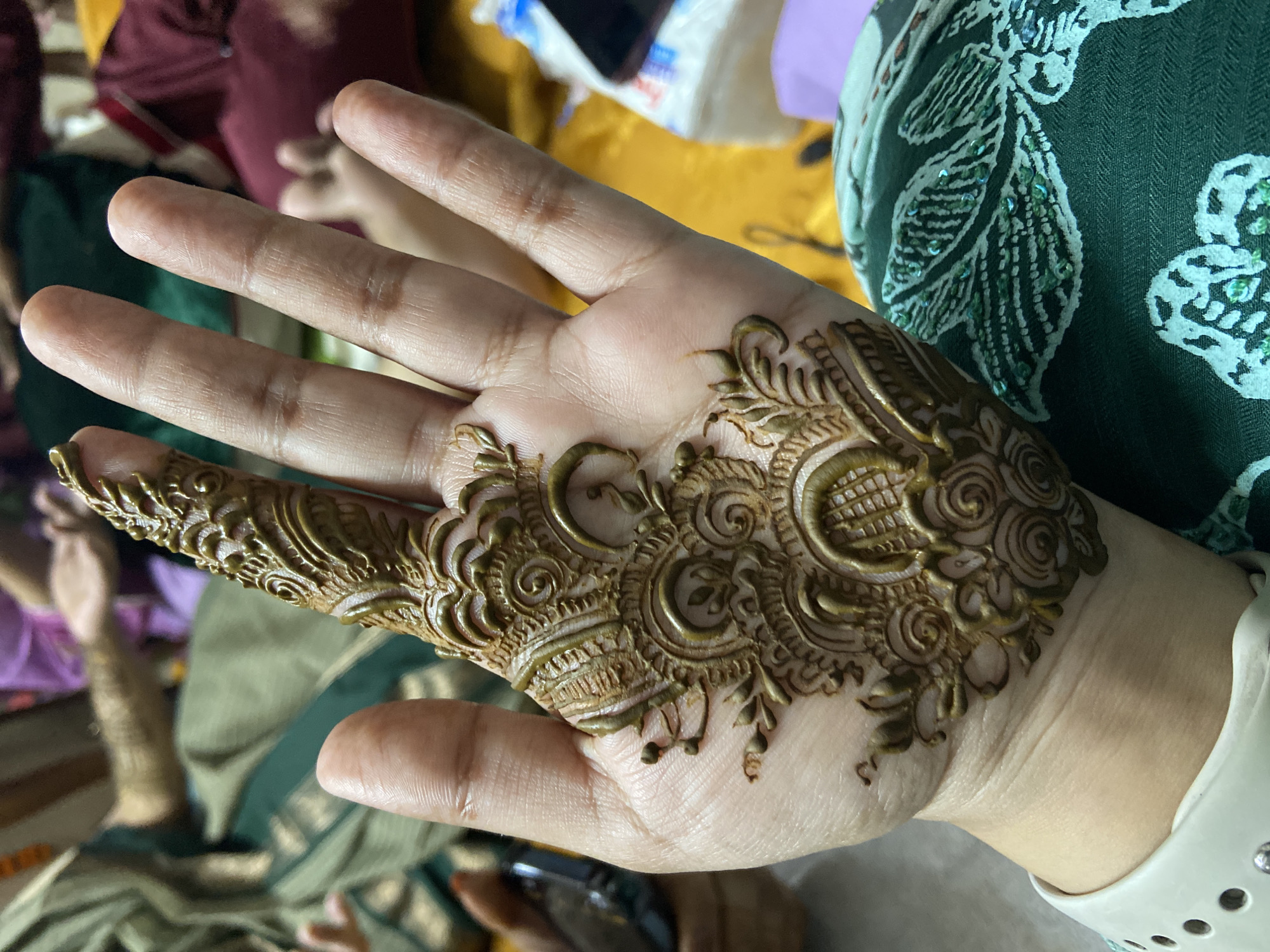 A close-up shot shows a right hand with elaborate henna designs covering the palm and index finger. The henna is a rich brown color with intricate floral and swirling patterns. The hand is light-skinned with visible veins. The fingers are slightly bent. A white smartwatch band is visible on the wrist.
The background is out of focus, but it appears to be an indoor setting with clothing and other objects scattered around. There are hints of green and yellow fabrics. The location is Dhaka, Bangladesh.