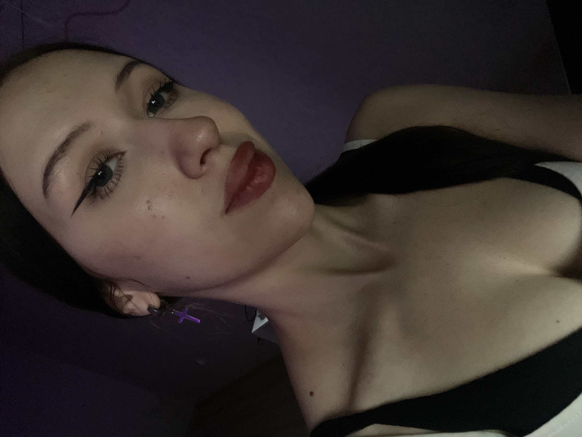 A young adult female is depicted in a close-up, chest-up portrait, looking downwards and slightly to her right, directly towards the camera, with a neutral expression. She has dark hair, which appears pulled back, and dark, well-defined eyebrows. Her eyes feature prominent black winged eyeliner and dark mascara on the eyelashes, with a visible light-colored iris. Her lips are full and coated with a glossy, reddish-brown lipstick. Several small moles or freckles are visible on her face and upper body, including one prominent mole on her left clavicle area.
She wears a dark-colored garment, likely black, with thin straps, layered over a white undershirt or bra, parts of which are visible at the neckline. A singular, bright purple, cross-shaped dangle earring is worn on her visible left earlobe, attached to a silver-colored hoop.
The setting appears to be indoors and dimly lit, with a dominant dark and indistinct background. A subtle purple ambient light emanates from the right side of the frame (viewer's perspective), highlighting parts of her face and shoulder. A faint, light-colored object, possibly furniture or equipment, is barely discernible in the dark background behind her left shoulder. The image provides no identifiable geographical markers, contextual elements, or specific objects that would allow for the determination of a location such as Votkinsk, Russia, or any other specific city/country.