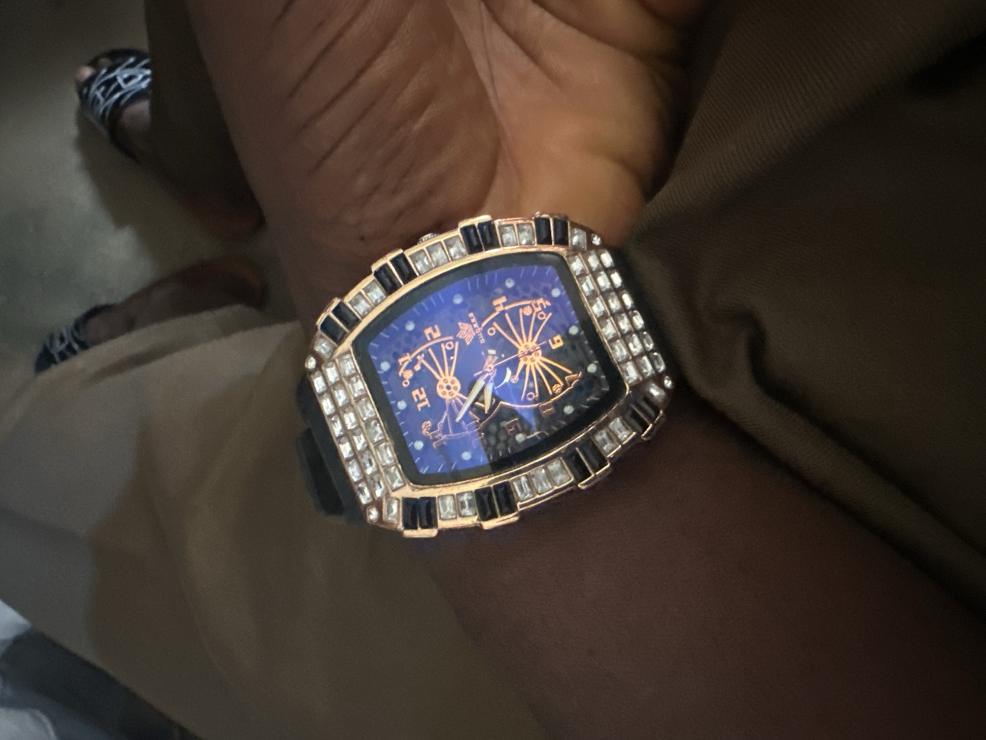 A dark-skinned individual is depicted sitting, with their left forearm extended towards the viewer, prominently displaying a watch. The watch features a tonneau-shaped, rose gold-toned case, heavily embellished with clear baguette-cut crystals along the upper and lower edges of the bezel, and square-cut crystals interspersed with black rectangular elements on the side bezels. The watch face is luminous blue, displaying an intricate gold-toned, starburst or gear-like design at its center. Orange-glowing Arabic numerals (12, 2, 4, 6, 8, 10) mark the hours, and gold-toned hands indicate the time. The brand name "D.U.B.K.E.R" is visible on the dial below the 12 o'clock position. The watch is fitted with a black rubber strap. The person is wearing light brown or khaki trousers, exhibiting visible folds and texture across the lap and leg. In the background, partially obscured and out of focus, a foot wearing a black slide sandal with white geometric patterns rests on a dark surface, possibly a floor. The scene is illuminated by ambient light, suggesting an indoor setting, and takes place in the city of Jos, Nigeria.
