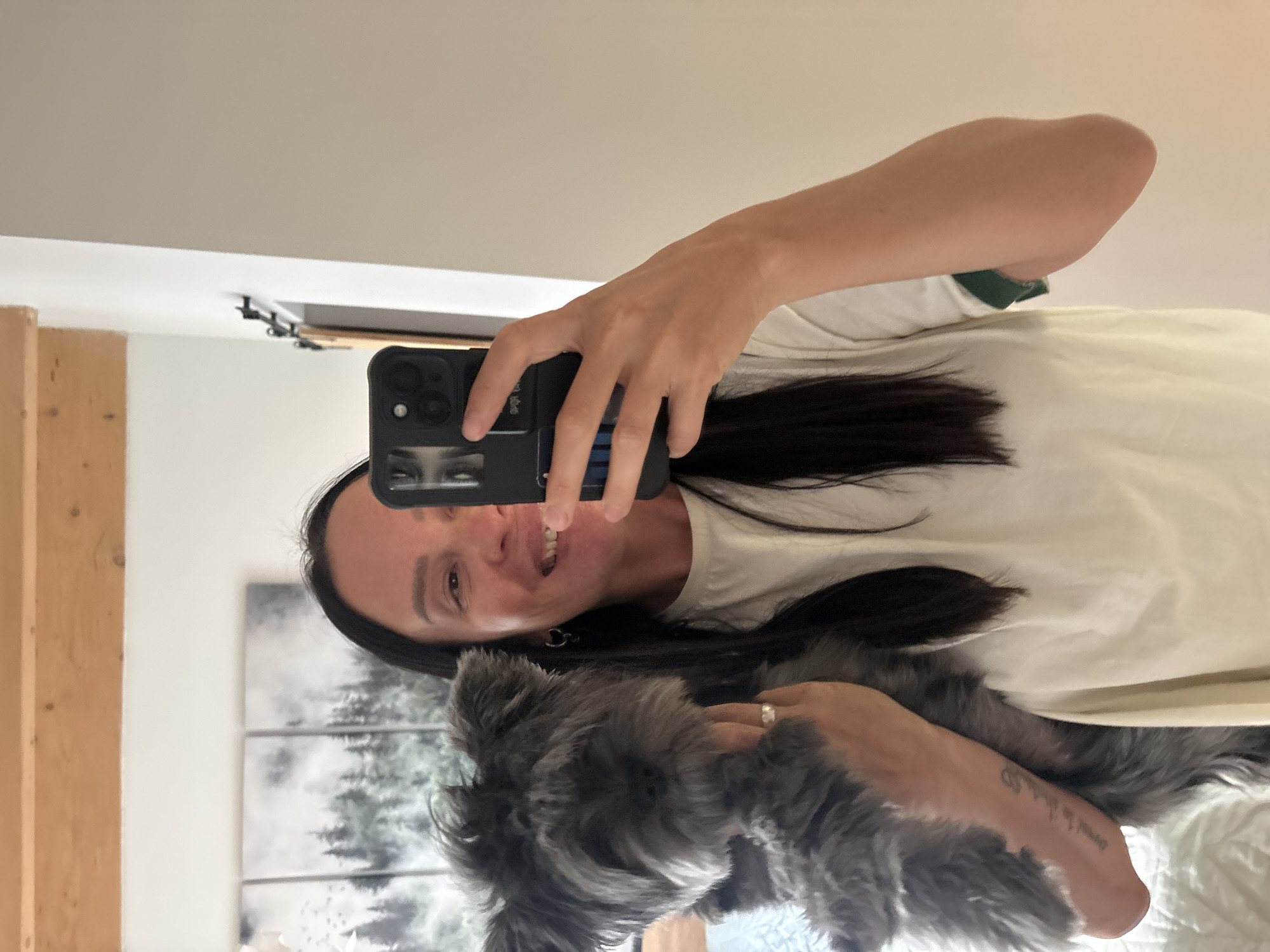 A woman is taking a selfie in a mirror, holding a small, gray dog. The woman has dark hair and is wearing a white t-shirt. She is holding a black smartphone. Behind her, a picture of a forest scene can be seen on the wall. There's a wooden beam visible on the left. The location is potentially an indoor space, but there are no specific details to indicate it is in Chaikovsky, Russia.