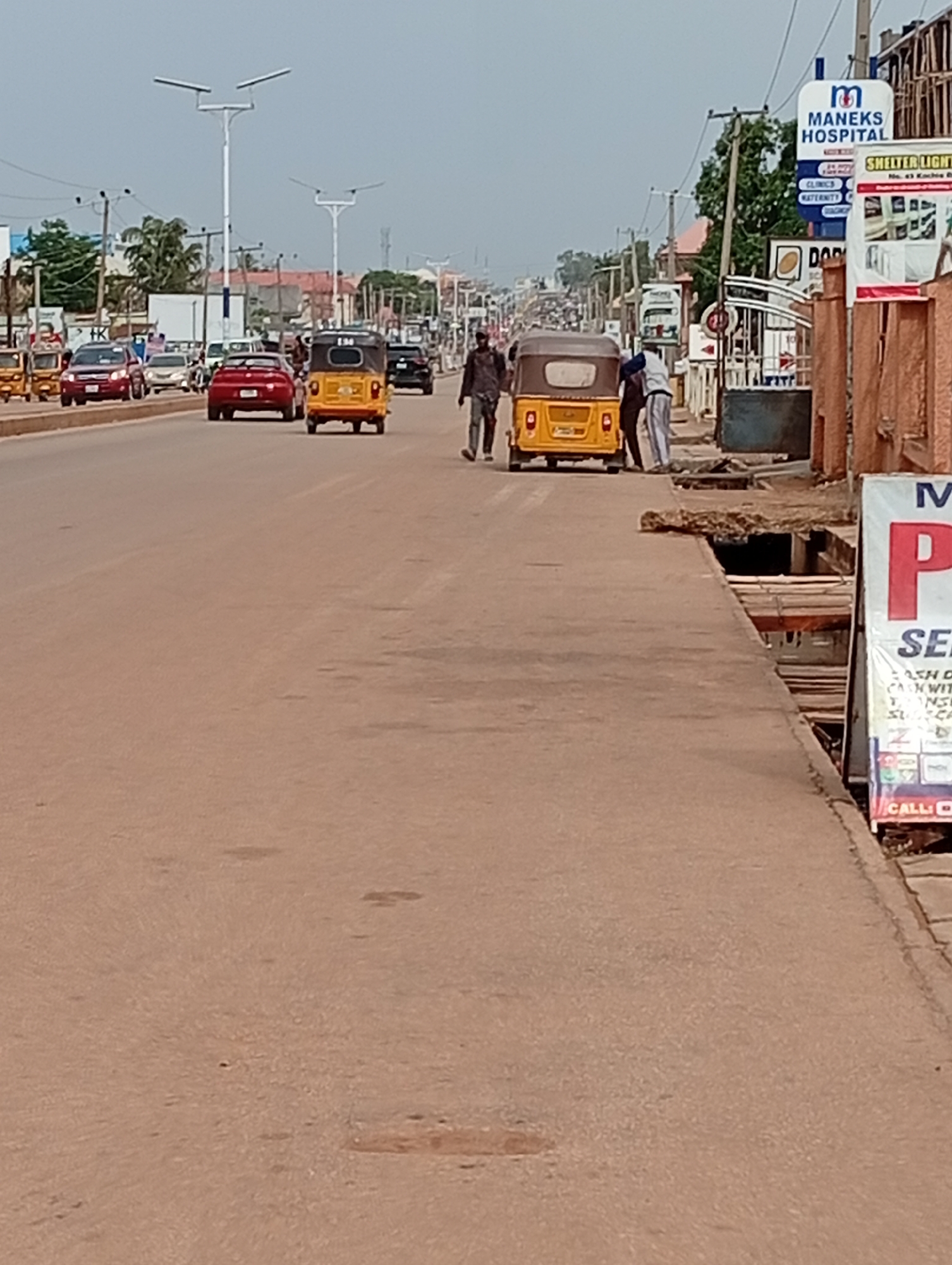 The image depicts a street scene in Sabon Tasha, Nigeria. A wide, tan-colored road extends into the distance with several cars and yellow three-wheeled vehicles (tuk-tuks) visible. Two tuk-tuks are closer to the camera: one is moving along the road and the other is being attended to by two men on the right side of the road near an open drain. A man in dark clothing is walking alongside the road. On the right side of the road, a sign for MANEKS HOSPITAL is visible, along with other commercial signs. Light poles line the street in the distance, and buildings are visible along the horizon.