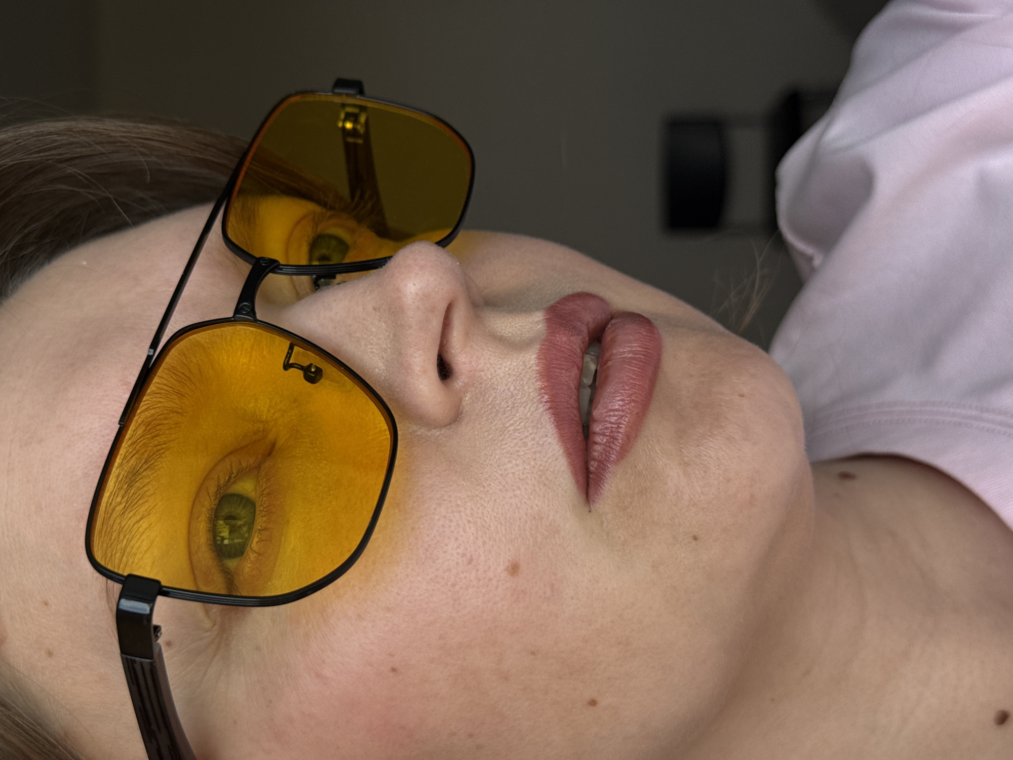 A close-up, angled shot depicts a person wearing yellow-tinted sunglasses. The individual is lying on their back, as indicated by the positioning of their head and the view. The reflection of a window is visible in the lenses of the sunglasses. The lips are colored with a matte lipstick. A shirt collar is visible on the right side of the image. The background is blurred. The location is not identifiable as Izhevsk, Russia from the provided image.