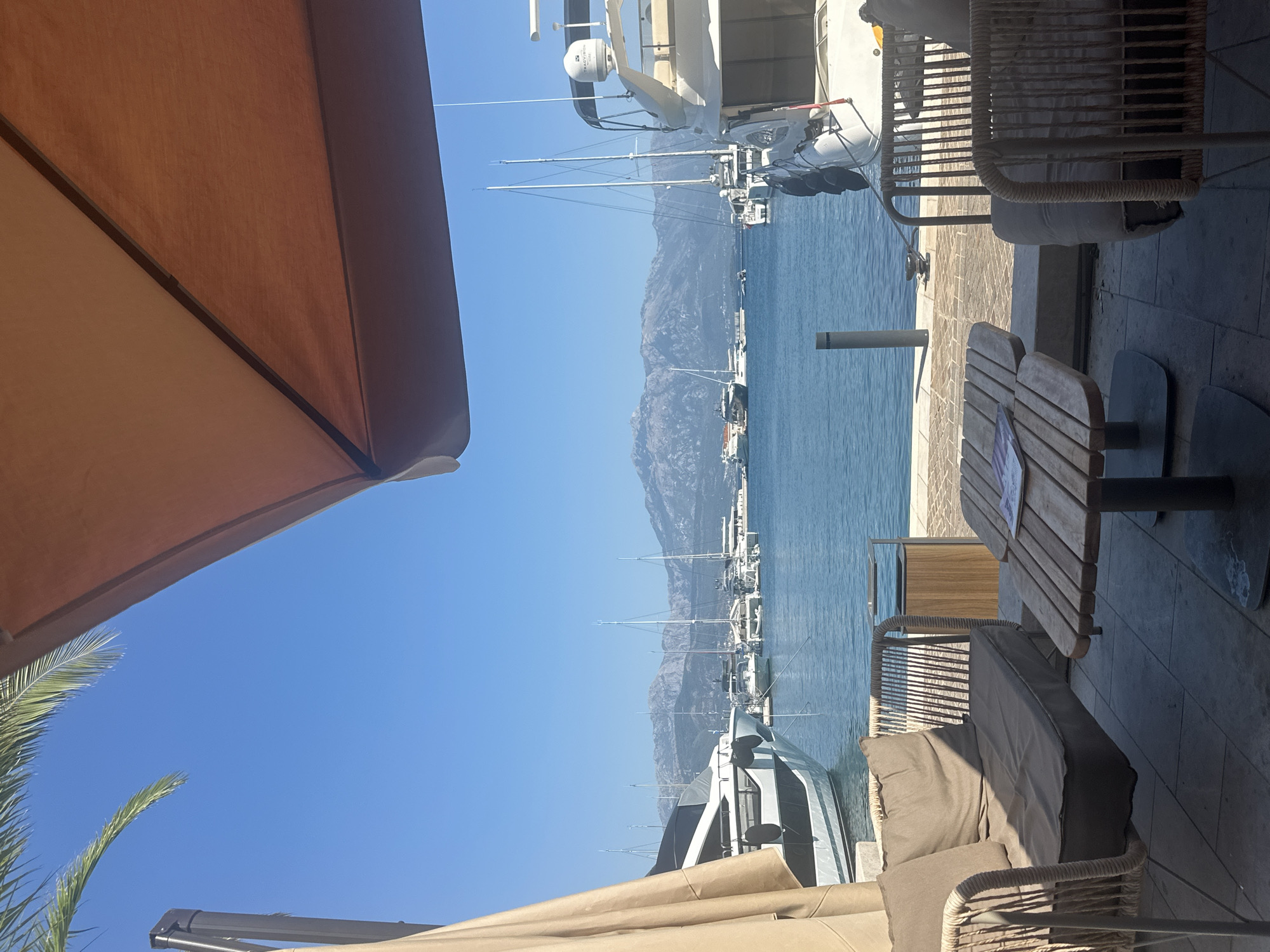 A casual outdoor seating area with a table and chairs is set up on a paved terrace overlooking a harbor filled with yachts. The scene is bathed in bright sunlight under a clear blue sky. In the foreground, a light brown upholstered sofa and a wooden coffee table with a magazine on it are visible. Beyond the seating area, the calm blue water of the harbor is dotted with numerous white yachts of various sizes, some with tall masts. In the distance, a range of rugged, light-brown mountains with some snow on their peaks forms the backdrop. The overall atmosphere suggests a serene and luxurious waterfront setting in Herceg Novi, Montenegro.