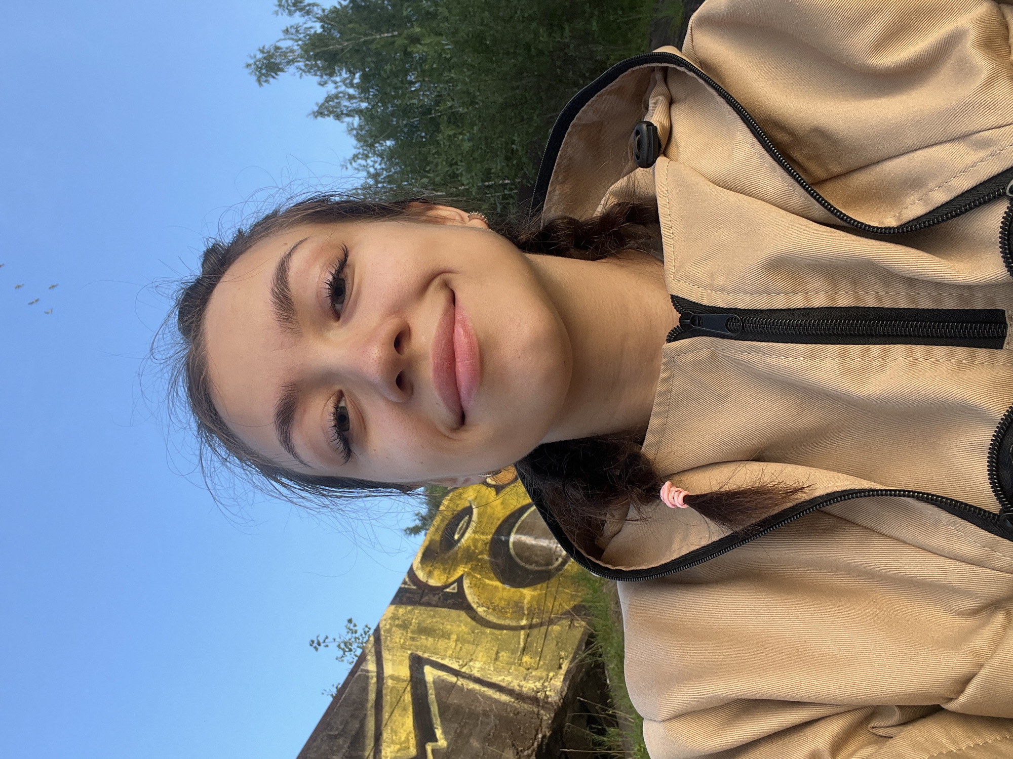 A young woman with brown hair in two braids, each tied with a pink elastic band, smiles at the camera. She is wearing a tan hooded jacket with a black zipper. In the background, a concrete structure with yellow graffiti is visible, along with trees and a clear blue sky with a few birds flying. The photograph was taken in Chernushka, Russia.