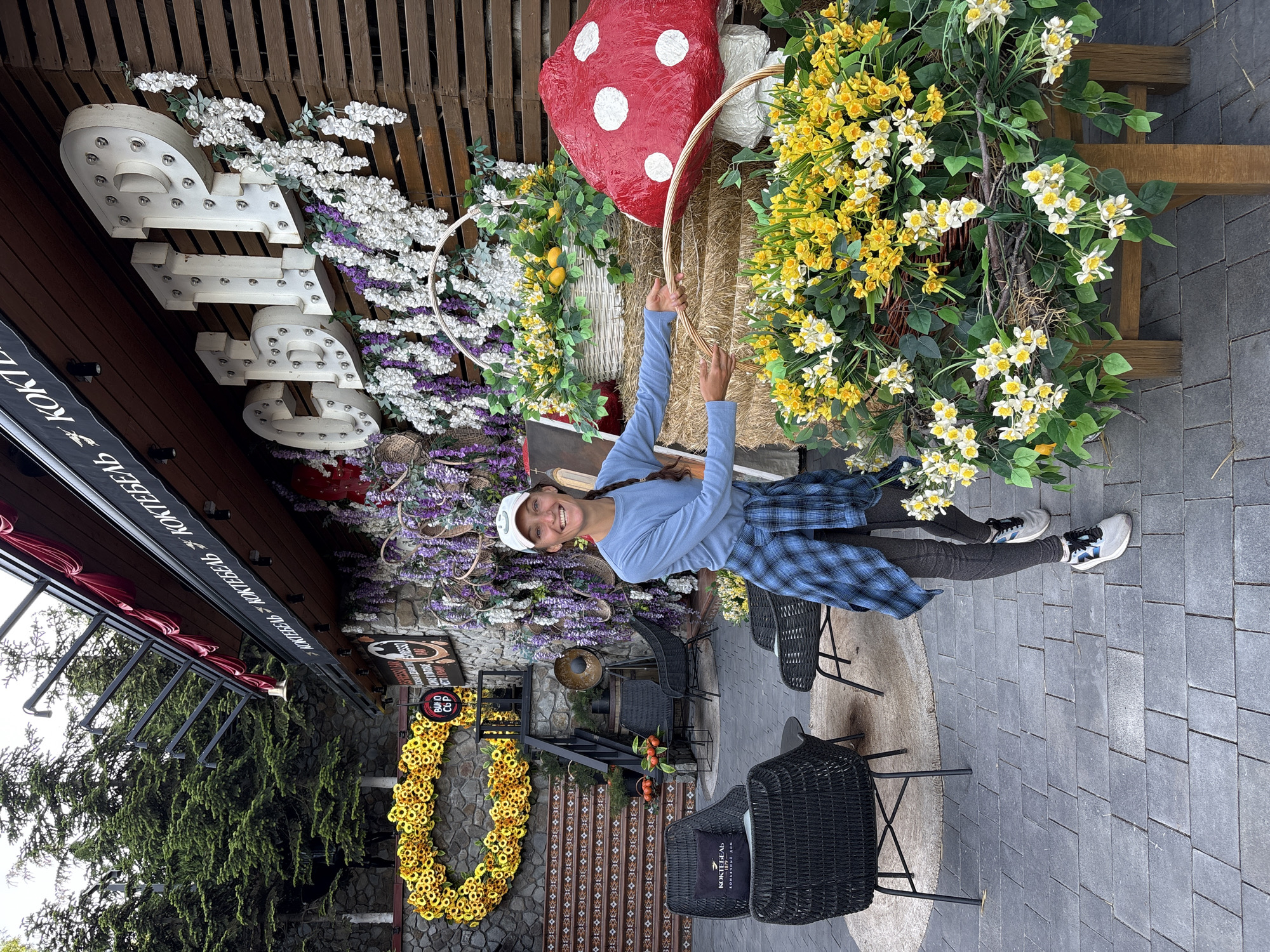 This image captures an outdoor scene at a decorated establishment in Kiparisnoe, Ukraine. A Caucasian woman stands smiling in the right-center, facing the viewer, wearing a white baseball cap, light blue long-sleeved shirt, dark grey leggings, and a blue and black plaid shirt tied around her waist. Her hair is braided and rests on her right shoulder. She holds a semi-circular wicker handle that is part of a multi-tiered artificial flower display.
The flower display features two hay bales, with a white wicker basket filled with artificial yellow and white flowers and green foliage, including two artificial lemons, resting on the upper bale. Below this, a larger arrangement of similar artificial yellow and white flowers is presented in a basket-like structure on a wooden stand. To the right of the display, a large red and white polka-dotted mushroom statue is visible.
Behind the woman and the flower display is the facade of a building, likely a cafe or bar, featuring dark wooden horizontal slats. Large white illuminated letters spelling "СБИР" (SBIR) or "СЫР" (SYR) are mounted on the facade and decorated with artificial white and purple wisteria-like flowers. Above the letters, a red awning is visible, bearing white Cyrillic text "КОКТЕБЕЛЬ" (Koktebel) repeated, separated by a cocktail glass icon. To the left of the main entrance, a stone wall is adorned with a large heart-shaped wreath made of yellow artificial sunflowers. Within this wreath is a circular sign reading "ВИНО" (Vino - Wine) and "СЫР" (Syr - Cheese). A set of tiled stairs with dark railings leads up past this display. An additional smaller sign on the building near the entrance reads "ESPRESSO BAR."
The foreground features an outdoor seating area with several dark wicker chairs and small tables on a paved grey stone surface. One chair includes a pillow with Cyrillic text "КОКТЕБЕЛЬ" and "КОНЬЯЧНЫЙ ДОМ" (Cognac House). Behind the stone wall and building, numerous green coniferous trees are visible under an overcast sky.
