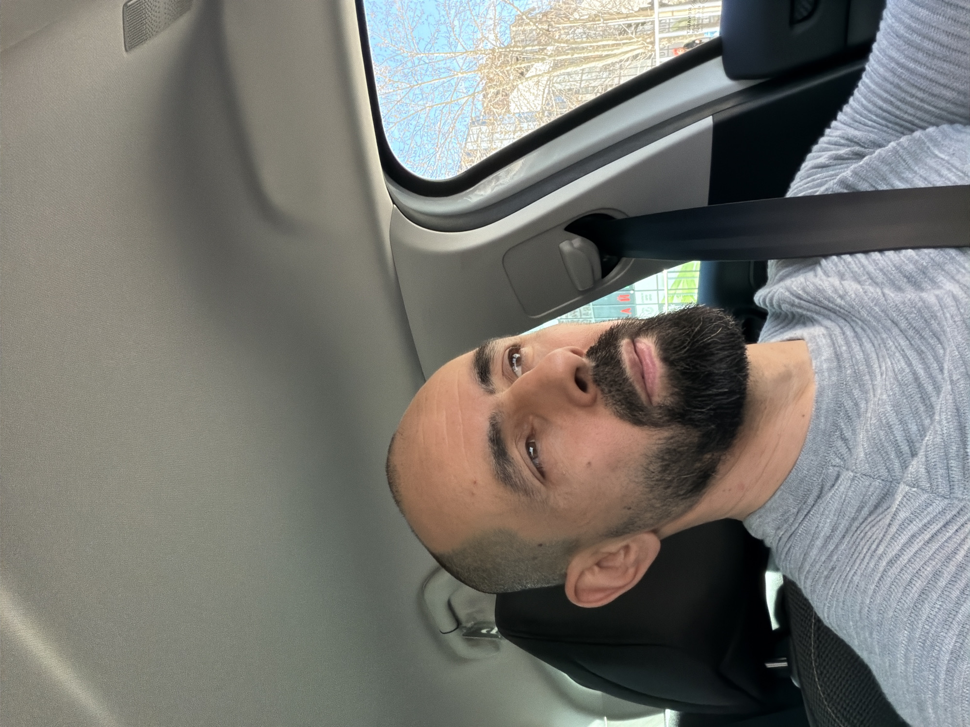 A man with a shaved head and a full beard is wearing a gray sweater and a seatbelt inside a car. He is looking upwards and to the left. Through the car window, a cityscape with bare trees and buildings can be seen. The exact location in Plovdiv, Bulgaria is not identifiable based on the visible background elements.