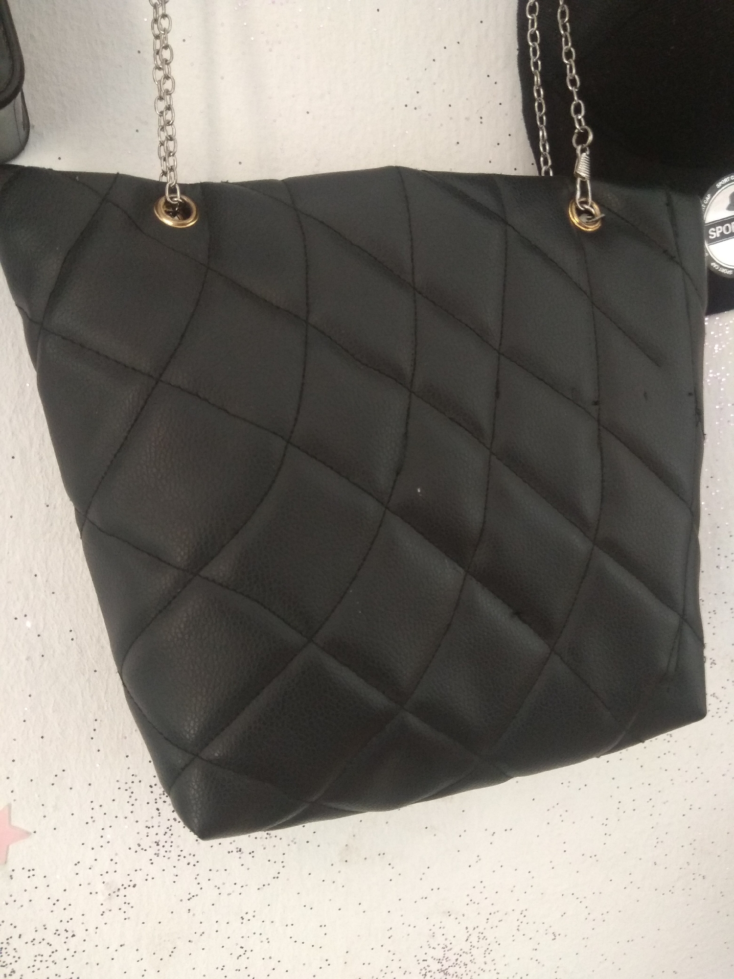 The image shows a close-up of a black quilted handbag with silver chain straps. The bag is the primary subject and is centrally positioned. The background is a textured white surface with scattered glitter, suggesting a decorative or festive setting. Part of a black hat with "SPORT CAP" visible in white text is also present on the right side of the frame. The lighting appears to be bright, possibly from an indoor setting. No people are visible in the image. The overall mood is neutral, focusing on the product itself. The location context provided is Jalingo, Nigeria, but there are no specific visual cues in the image to confirm this location.