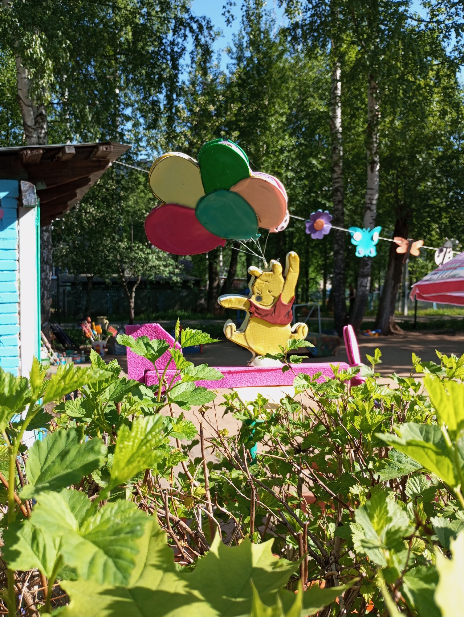 The image shows a brightly colored outdoor scene, possibly a playground or recreational area in Izhevsk, Russia. In the foreground is lush green foliage, partially obscuring a pink picnic table. Atop the table sits a cutout figure of Winnie the Pooh holding a bunch of balloons colored yellow, green, and pink. A string of decorative cutouts, including flowers and butterflies, stretches in the background. The left side of the frame shows a section of a small blue building, while a striped umbrella can be seen on the right. The background features tall trees under a clear blue sky.
