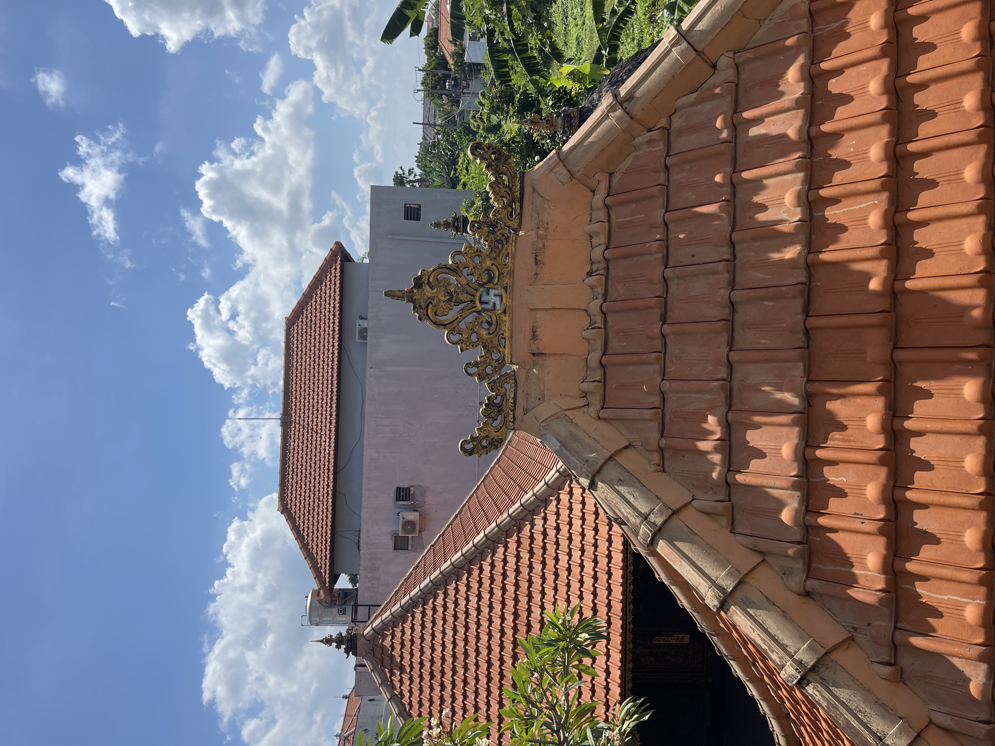 The image captures a rooftop view in Kuta, Indonesia, under a bright blue sky with scattered white clouds. The foreground is dominated by terracotta roof tiles, some appearing aged and slightly weathered. A decorative golden element, possibly a crest or finial with intricate scrollwork and a swastika symbol, sits atop one of the tiled roofs. Behind this, another tiled roof extends, and further in the background, a light pink building with visible air conditioning units and windows is present. Beyond the buildings, lush green vegetation, including banana leaves, is abundant. The scene suggests a typical residential or temple area in Kuta, characterized by traditional roofing and tropical foliage. No people are visible in the image.