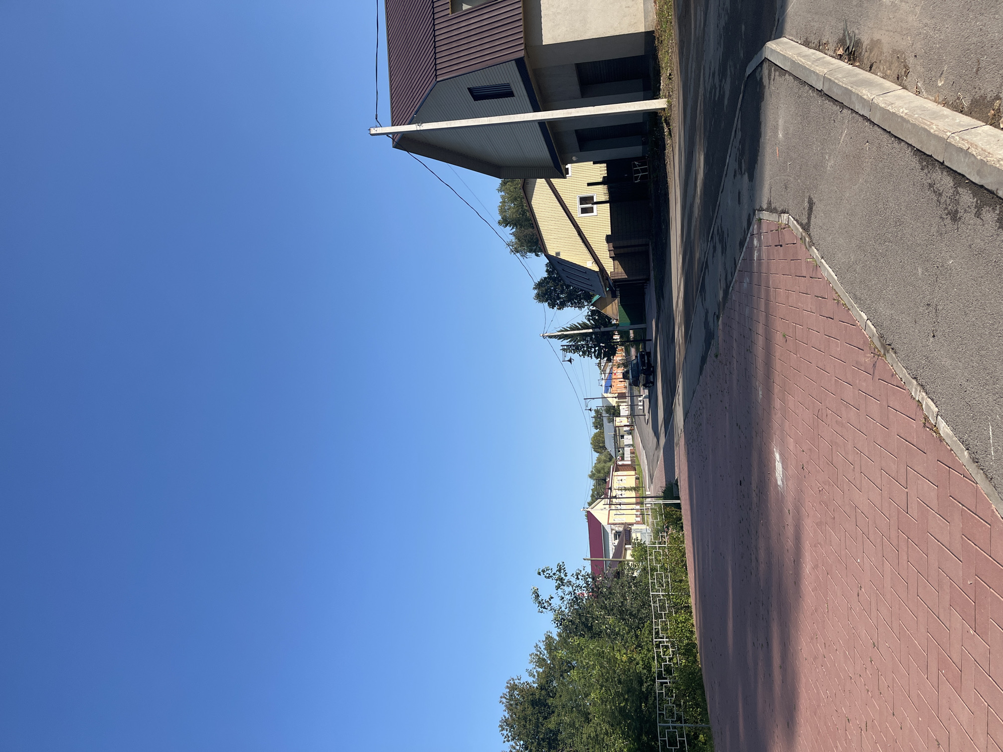 The image shows a street scene in Ufa, Russia, on a clear, sunny day. The foreground is dominated by a red brick paved area, likely a sidewalk or plaza, with a concrete curb. To the right of this is a narrow asphalt path, also bordered by a concrete curb. The street curves to the left in the midground, lined with several buildings. On the right side of the street, a large building with a light-colored facade and a dark, sloping roof is prominent. Further down the street, other buildings with varying architectural styles and colors are visible, some with red or yellow roofs. Trees and foliage are present on the left side of the image, bordering the brick-paved area. A white metal fence runs along the edge of the foliage. A few streetlights can be seen along the street. A black car is parked on the street in the midground. The sky is a clear, bright blue. The scene appears to be a quiet residential or commercial street.