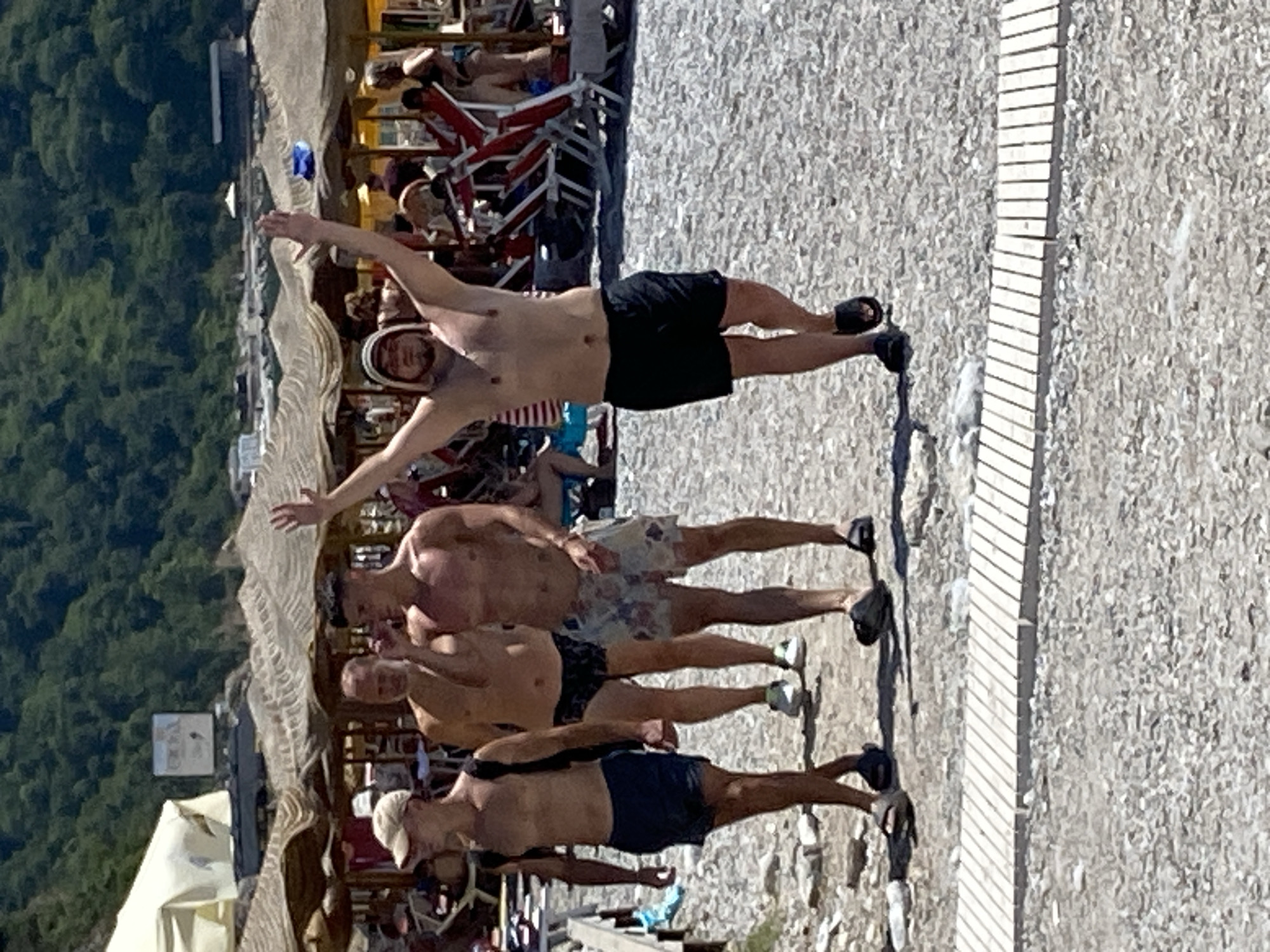 The image captures a sunny day at a beach in Prijevor, Montenegro. Four men are walking on a pebble beach. The man in the foreground on the right is wearing black shorts and has his arms raised in the air, his head covered by a patterned cloth. To his left, another man wears floral-patterned shorts and walks with his arms at his sides. Further left, a man in black swim trunks and a man in blue shorts and a cap are walking together. In the background, beach umbrellas and lounge chairs are visible, with a lush green mountainside rising behind them. The sky is bright and clear, indicating fair weather.