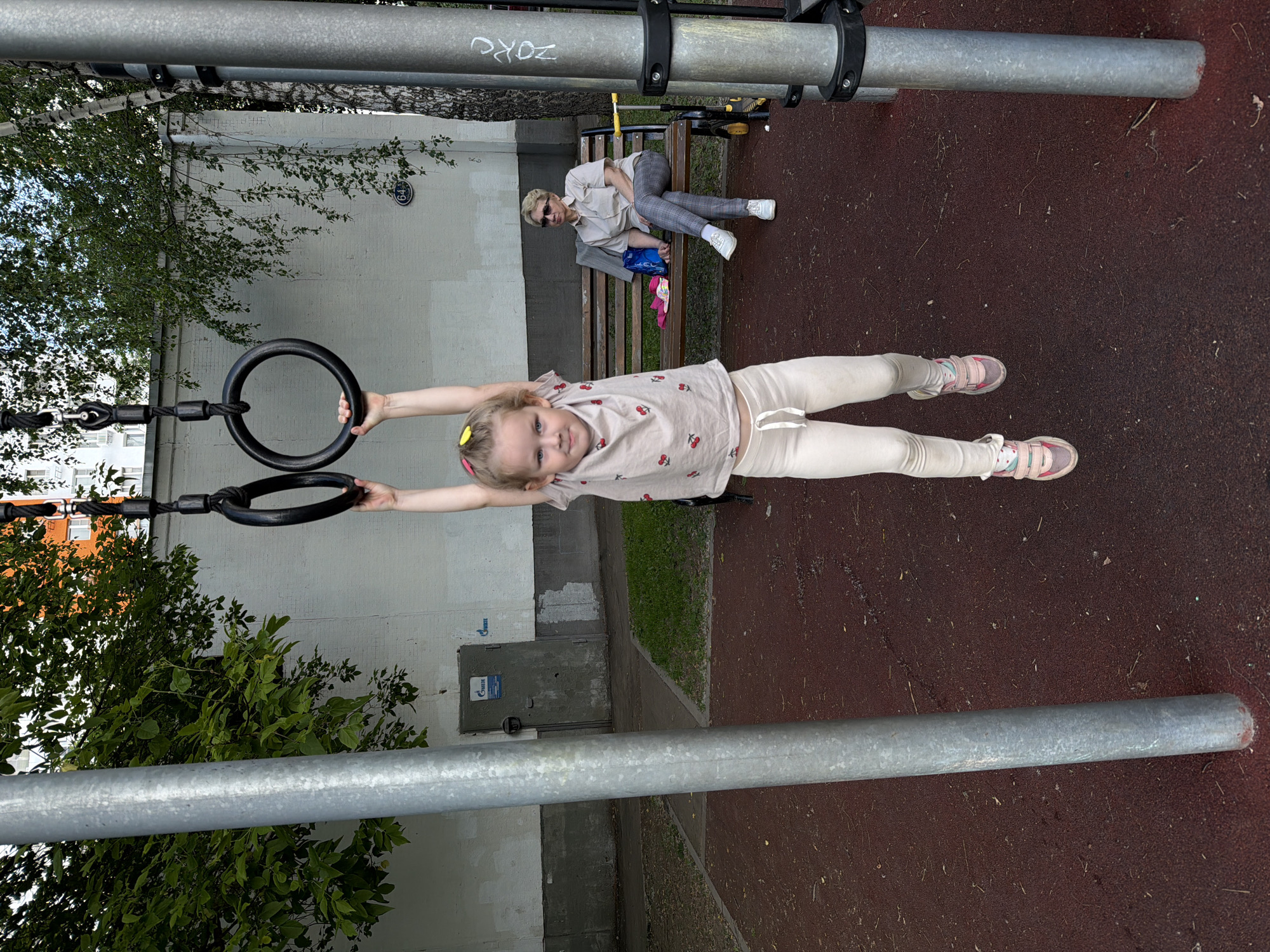 This image captures a bright daytime scene at an outdoor playground in Khimki, Russia, featuring a young girl and an adult woman.
In the foreground, a young girl with light hair and a yellow hair clip is actively engaged, hanging horizontally from a set of black gymnastic rings. She wears a light-colored t-shirt adorned with red cherry prints, light leggings, and pink and white sneakers. Her arms are outstretched, maintaining her balance as she looks towards the viewer with a composed expression.
To the right and slightly behind the girl, an adult woman sits relaxed on a wooden bench. She is dressed in a light-colored collared shirt, plaid trousers, and white sneakers, with sunglasses resting on her face. A blue bag and a pink hat are placed on the bench next to her.
The playground surface is a reddish-brown, textured material, likely rubber mulch, providing a soft landing area. Overhead, sturdy metal horizontal bars are visible, one of which has graffiti-like text "ZORG" or similar inscribed on it. In the background, a light-colored building is partially obscured by lush green trees and foliage. Notable details on the building include a circular sign with the number "64" and a blue sign featuring the "GAZPROM" logo on a door or panel. The atmosphere appears calm and pleasant, suitable for outdoor activities.