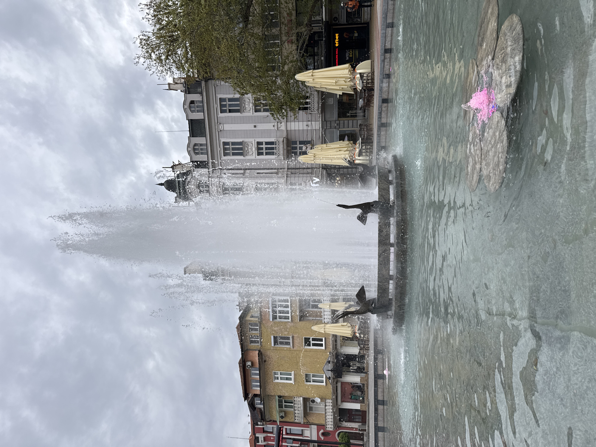 A vibrant public square or pedestrian area in Plovdiv, Bulgaria, is captured in this image, featuring a large, active fountain under an overcast sky. The scene is depicted from an eye-level perspective, looking across the fountain towards a row of buildings.
In the foreground, a multi-tiered fountain dominates the central and lower portions of the frame. A powerful, tall jet of water sprays high into the air from the fountain's center, creating a misty effect. Smaller water features are also visible, including a decorative element shaped like a flower with a distinct pink light emanating from its water jet, positioned towards the right side of the fountain basin. Dark-colored, abstract sculptural elements resembling birds are situated along the fountain's edge.
Behind the fountain, a diverse collection of multi-story buildings lines the square. On the left, a light-grey building with classic architectural details, including arched windows and ornate facades, stands prominently. To its right, a mustard-yellow building with numerous rectangular windows and visible air conditioning units adds a contrasting aesthetic. Further along, a building with a dark facade features a clearly visible sign displaying "efbet," a Bulgarian betting company, with additional text "WELCO BO" likely part of "WELCOME BONUS" below it. Several outdoor cafes or restaurants are suggested by the presence of numerous beige/light yellow parasols and accompanying tables and chairs, indicating an area for public gathering and leisure. Green foliage from trees is visible above and between some of the buildings, adding a natural element to the urban setting.
The sky is uniformly overcast with soft, grey clouds, suggesting a cloudy daytime scene without direct sunlight. A few indistinct figures of people can be seen in the background, walking or near the buildings, but they are not central subjects. The image conveys the everyday atmosphere of a bustling city center.