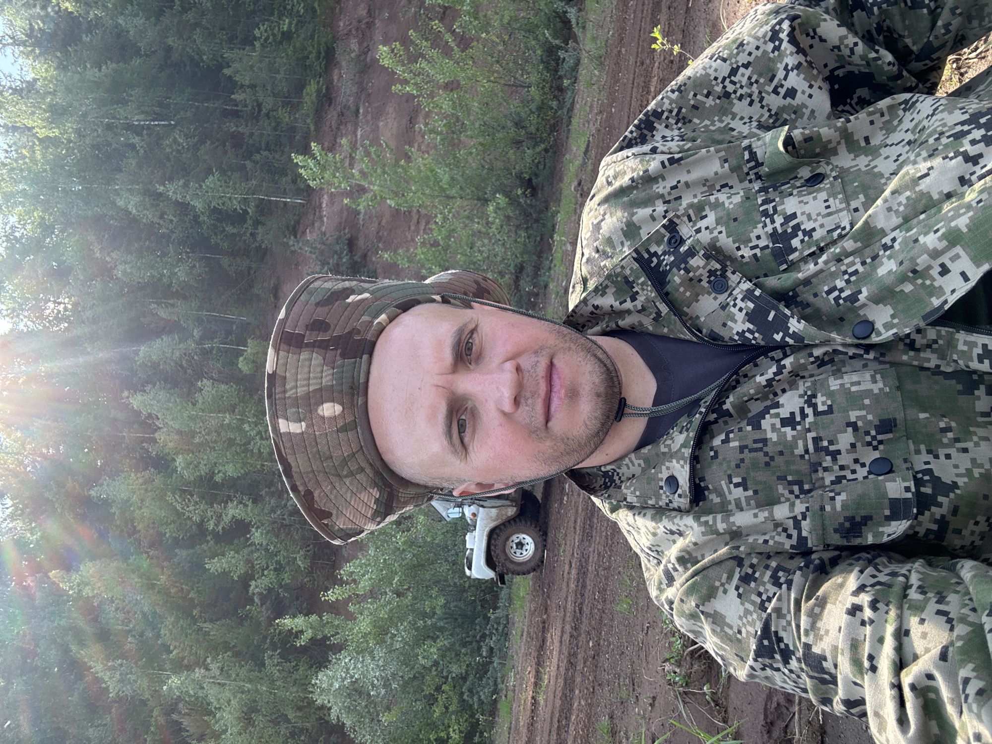 A man in camouflage clothing and a boonie hat stands in the foreground, looking at the camera. Behind him, a white off-road vehicle is parked on a dirt road. The background consists of a dense forest on a hillside, with sunlight filtering through the trees. The scene appears to be in a rural or natural setting, possibly in the Прикамский region of Russia. The overall impression is of an outdoor adventure or recreational activity.
