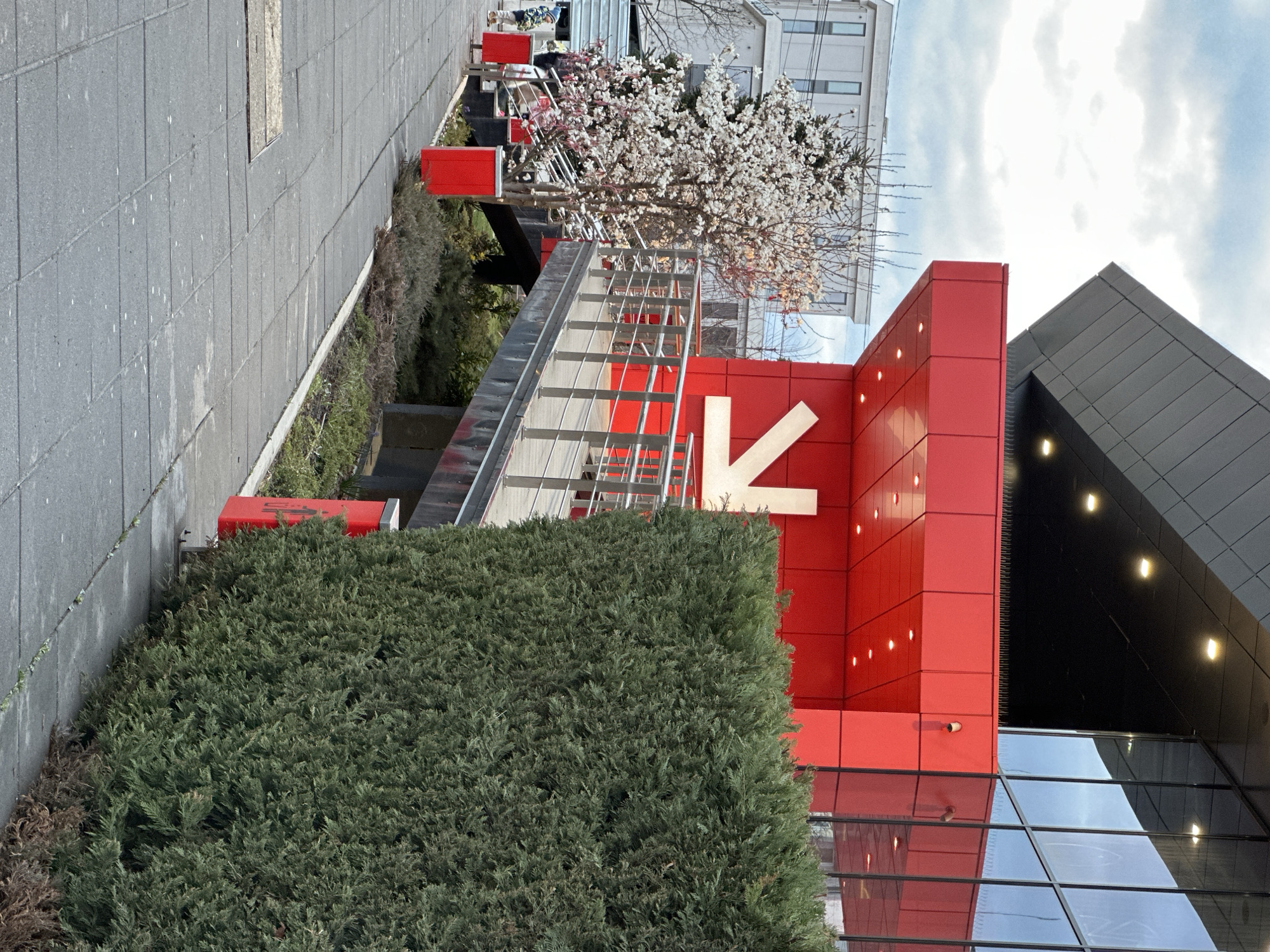 The image shows an outdoor scene in Sofia, Bulgaria, featuring an urban landscape with architectural elements. The main focus is a modern building with a red facade and a large white arrow pointing to the left. The building also features a glass facade, reflecting the surroundings. A staircase with metal railings leads towards the building. Lush green bushes partially obscure the view. A blossoming tree adds a touch of nature to the scene. The sky is partly cloudy. The scene is well-lit, suggesting it is daytime.