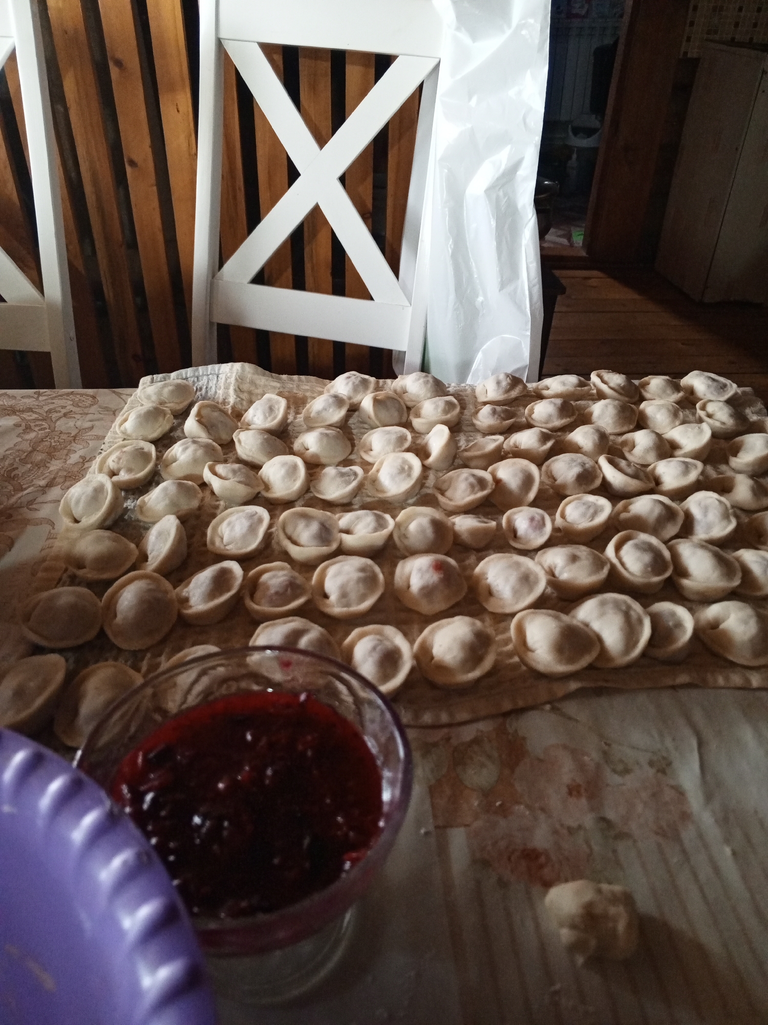 In a domestic setting in Petrovka, Russia, a close-up shot focuses on a table covered with a patterned tablecloth, upon which numerous uncooked pelmeni (dumplings) are arranged in rows. The pelmeni, made from white dough and containing a filling, are spread across a light-colored textured cloth. In the lower left of the frame, a translucent glass bowl holds a dark red jam or compote, likely a condiment for the dumplings. Partially visible in the foreground is a purple ridged plate. In the background, the image shows the lower portion of two white wooden chairs with a distinctive cross-shaped design on their backs, positioned against a backdrop of wooden planks. A large white plastic sheet hangs to the right of the chairs. The overall scene suggests food preparation in a home environment.