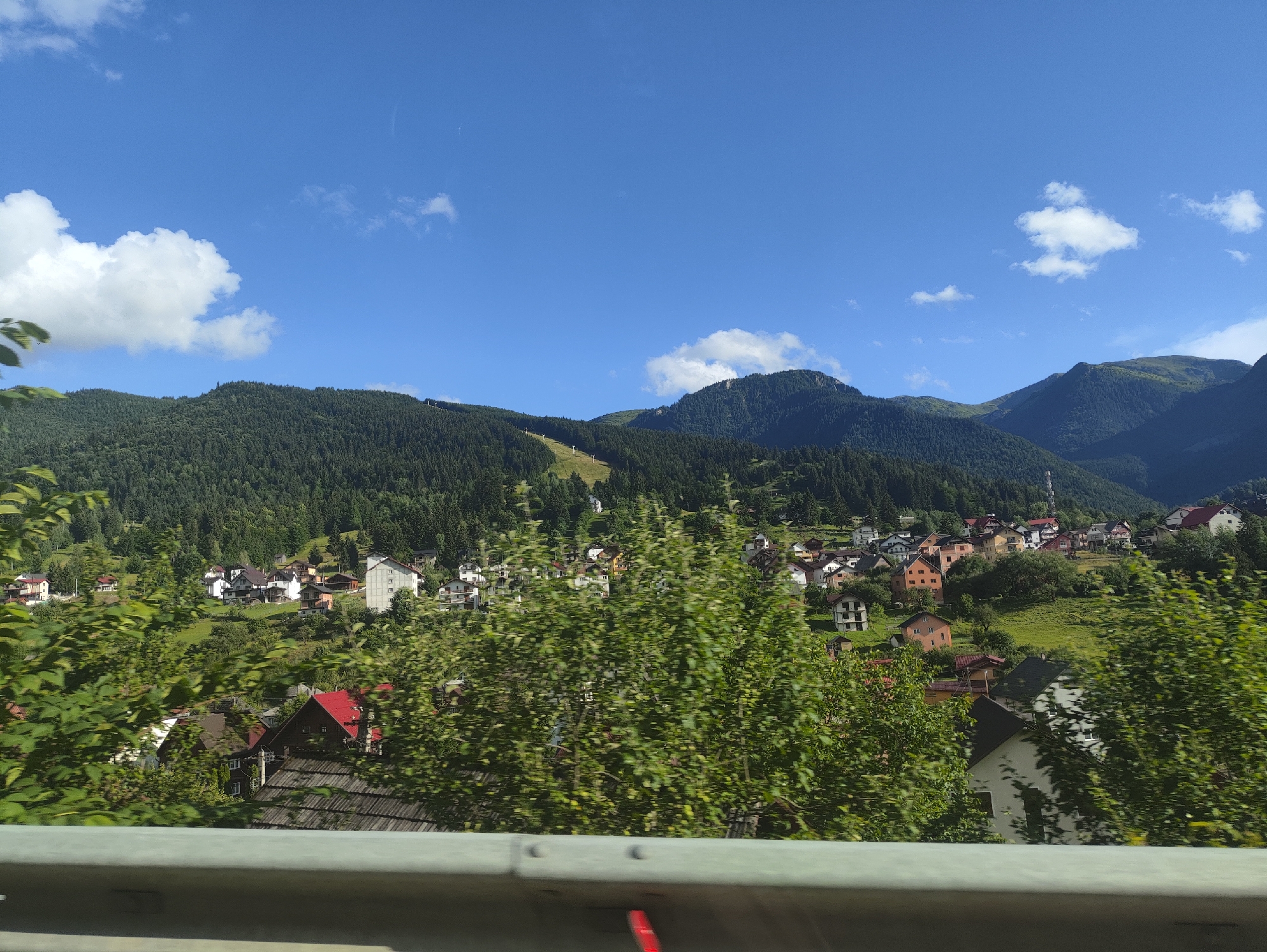 The image depicts a view of the city of Borșa, Romania, under a clear blue sky with scattered white clouds. In the foreground, a blurred section of a grey metal guardrail runs horizontally across the bottom of the frame. Behind the guardrail, lush green foliage from trees and bushes obscures a portion of the scene, some of which appears slightly motion-blurred.
Further into the scene, a hillside town is visible, nestled amongst rolling green hills and dense evergreen forests that cover the surrounding mountains. The town consists of numerous houses with red, brown, and grey roofs, showcasing a mix of architectural styles. Some houses are brightly colored in orange and yellow, while others are painted white or have darker facades. A ski slope is discernible on one of the forested mountainsides, indicating the area's potential for winter sports. The overall impression is that of a picturesque mountain village on a sunny day.