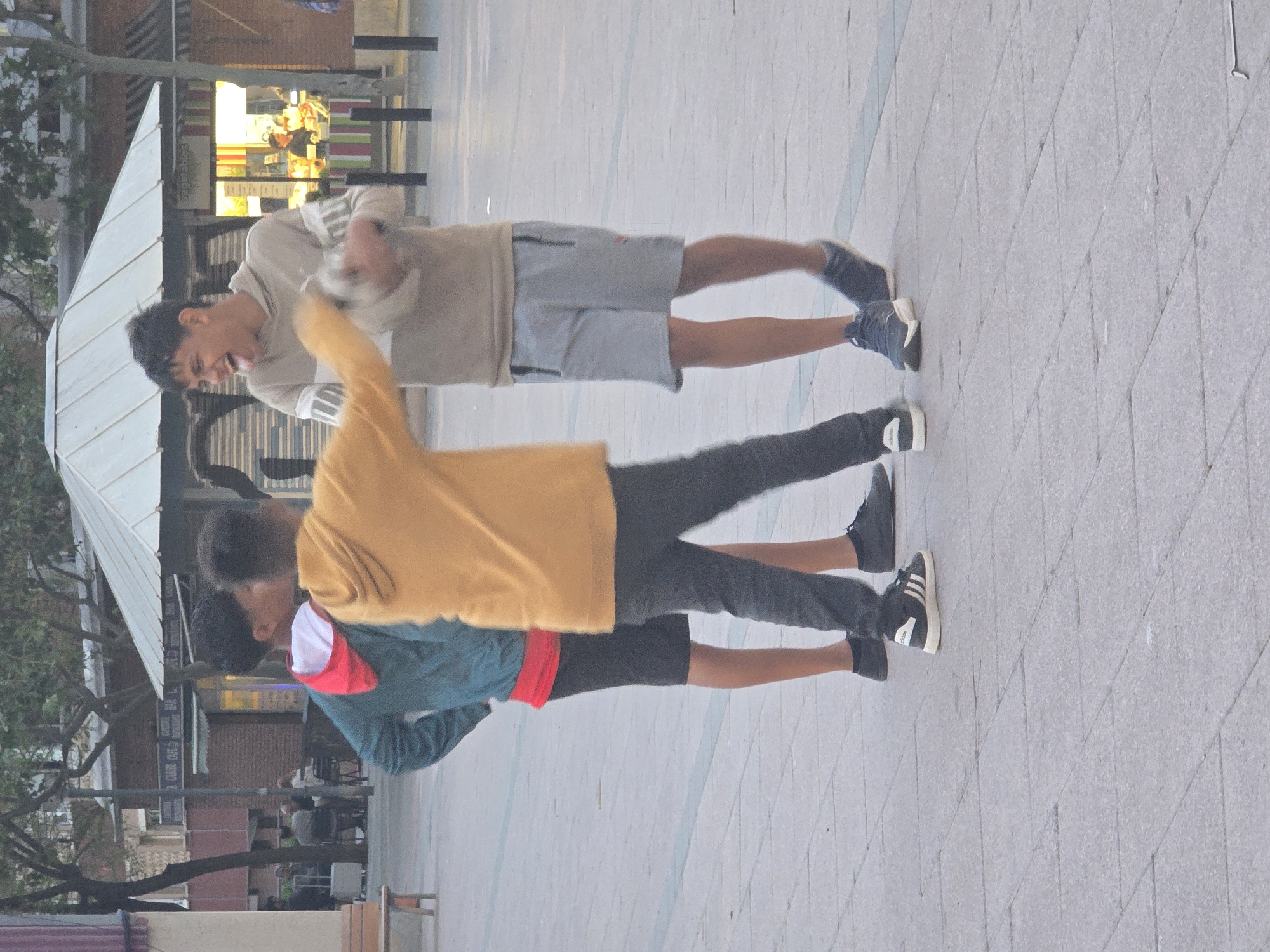 Two individuals stand on a paved walkway in Cornellà De Llobregat, Spain. The first person, positioned at the top of the image, wears a beige hoodie and grey shorts, with dark-colored sneakers on their feet. They are sticking out their tongue and appearing to be laughing. The second person is below them. The second person wears a yellow sweatshirt over a teal and red hoodie, dark trousers and dark sneakers with white stripes. A building is visible on the left, partially obscured by trees. A storefront with striped awnings and transparent panels is behind the people. The pavement is light grey with a geometrical pattern.