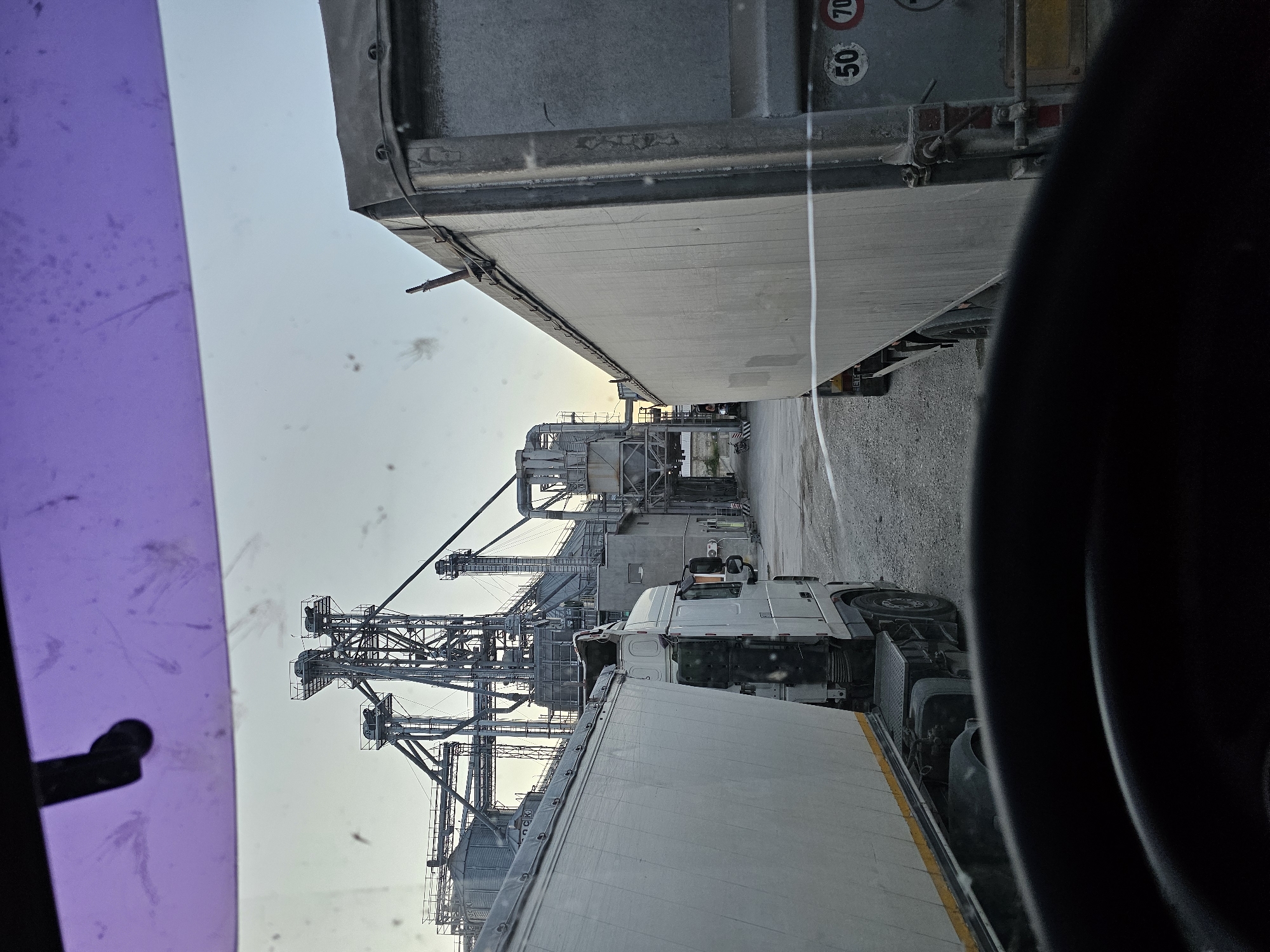 The image shows a view from inside a truck cab, looking out at an industrial facility. Two large white semi-trailers are parked side-by-side, with the cab of the truck on the left partially visible. In the background, a complex of grain silos and processing equipment rises against a clear sky. The ground is covered in gravel. There are no people visible in the scene. The overall impression is one of an industrial or agricultural logistical hub. The presence of speed limit signs (50 and 70) on the trailer on the right suggests a road within the facility or its immediate vicinity. The purple tint at the top of the image is from the truck's windshield or a sun visor. The image is taken during daylight hours, possibly late afternoon or early morning due to the soft lighting. There is no information in the image to indicate the location is Sofia, Moldova.
