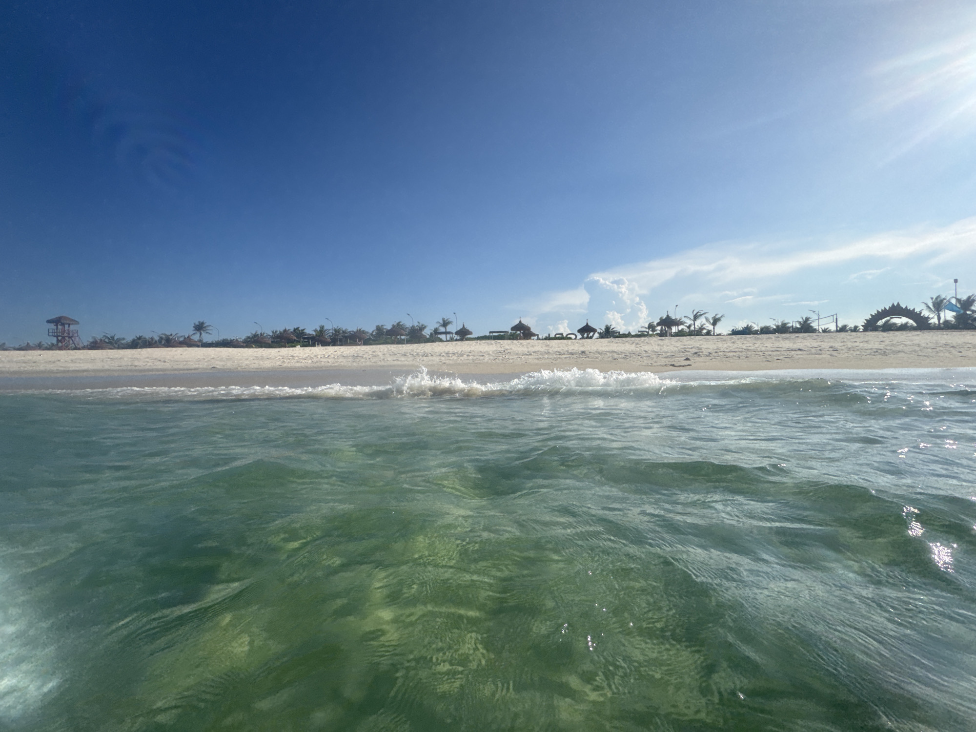 The image depicts a serene beach scene in Bac Cam Ranh Ward, Vietnam, on a clear, sunny day. The foreground is dominated by the crystal-clear, green-tinted ocean water, with gentle waves lapping at the shore. The seabed is visible through the translucent water, with sandy patches and subtle ripples. In the middle ground, a strip of pristine white sand forms the beach, leading to the horizon. Along the shoreline, several thatched-roof cabanas and palm trees are scattered, suggesting a resort or public beach area. Further in the distance, a red lifeguard tower stands prominently, indicating a monitored area. The sky is a vibrant, cloudless blue, with the sun casting a bright glare on the right side of the frame. The overall impression is one of tranquility and a picturesque tropical getaway.
