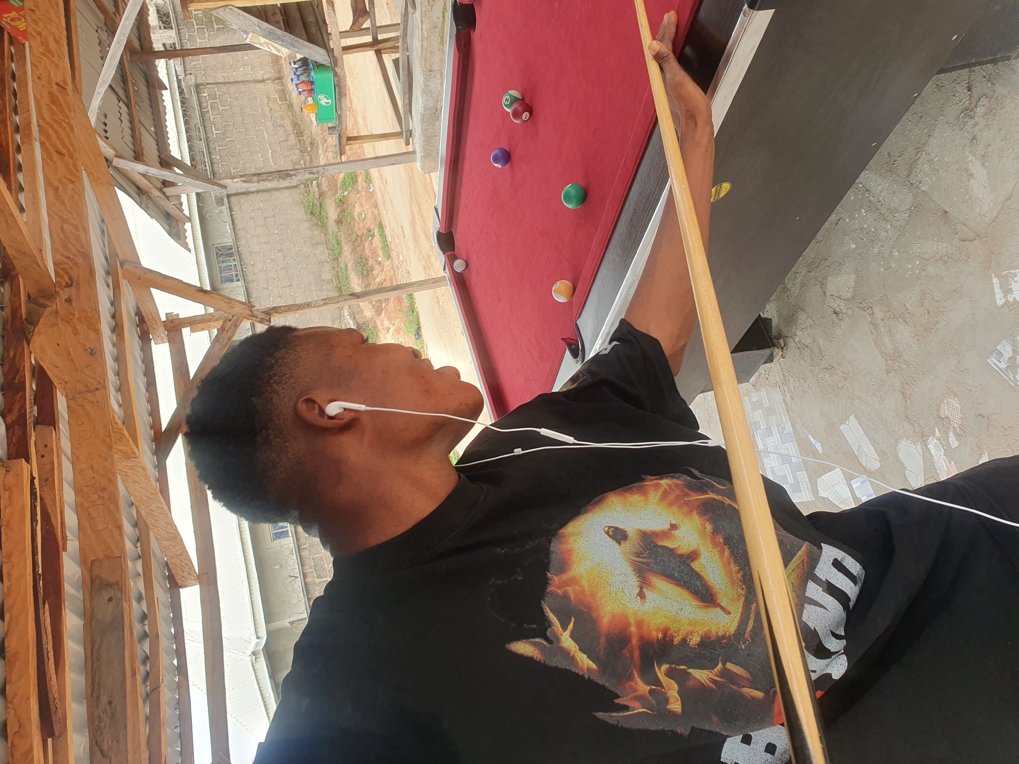 A Black male wearing a black t-shirt with a graphic of Jesus on it and white earbuds is playing pool on a red felt table under a rustic wooden shelter. He is holding a pool cue with his right hand, aiming at a white ball on the table. There are several other pool balls scattered on the table, including a purple ball, a yellow ball, a green ball, and two striped balls, one red and white, and the other brown and white. The background shows a wall made of concrete blocks, a fence, and some vegetation. The scene appears to be outdoors, likely in Lokoja, Nigeria, given the context.