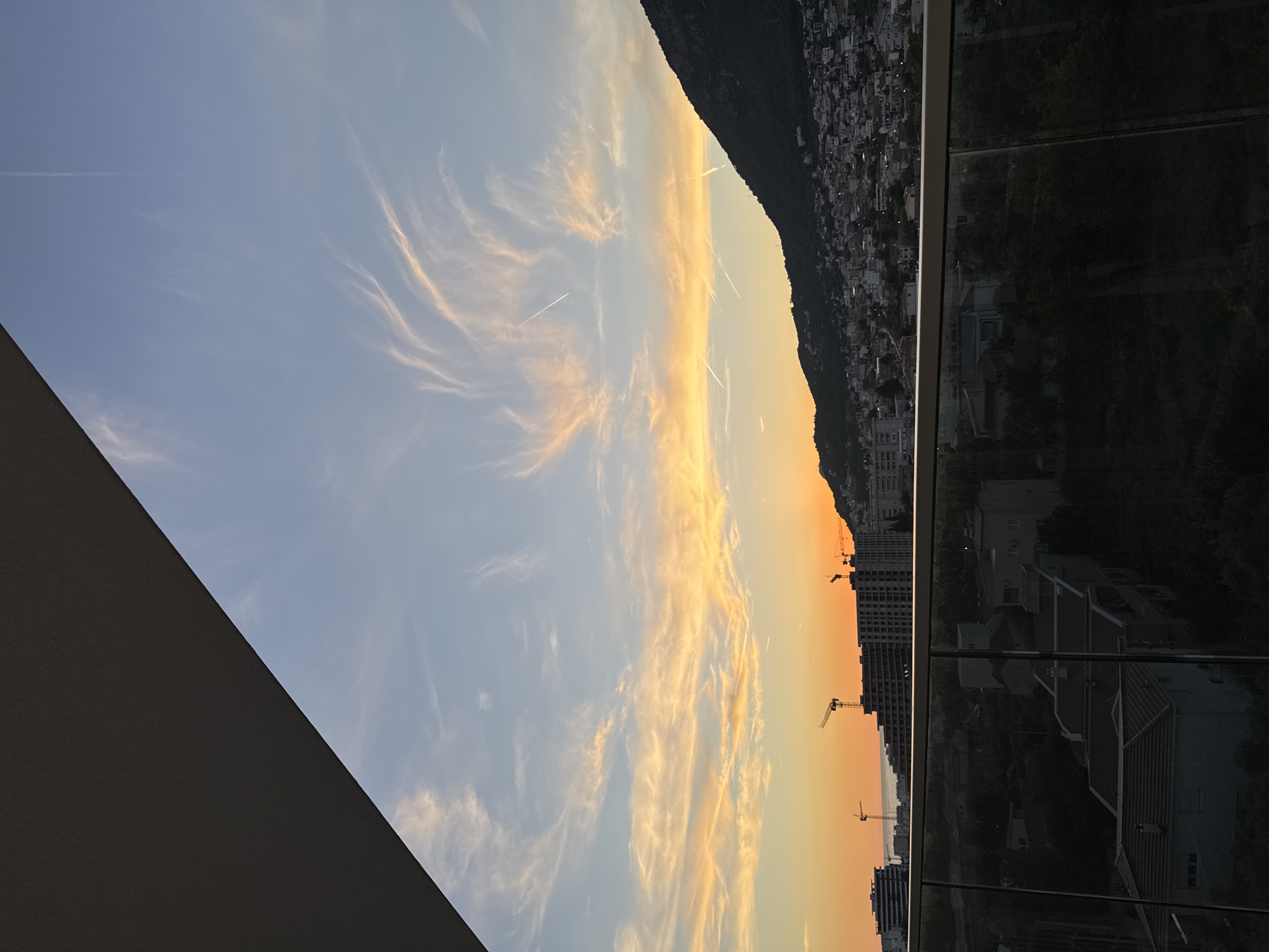 An aerial view of a city with buildings and a mountain range behind it under a colorful sunset sky. The city is nestled on a hillside, with many buildings of varying sizes and styles visible. A large, dark mountain looms over the city, its peak shrouded in shadow. The sky is a vibrant mix of orange, yellow, and blue, with wispy clouds scattered across it. The sun appears to be setting behind the mountain, casting a warm glow over the scene. In the foreground, a balcony railing made of glass and metal is visible, suggesting the photo was taken from a building overlooking the city. The overall impression is one of a serene and picturesque sunset over a coastal city. It is likely Šušanj, Montenegro given the hilly terrain and coastal proximity.