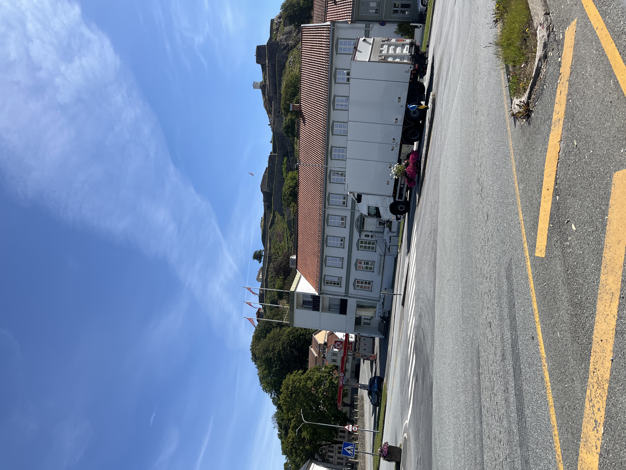 The image captures a sunlit daytime scene in the city of Halden, Norway, focusing on a section of road and buildings leading up to the imposing Fredriksten Fortress. The foreground shows a paved road with prominent yellow directional markings and a pedestrian crossing. To the left, a blue car is parked near a gas station with a red "Circle K" sign. Behind the gas station, several residential buildings with red tile roofs are visible. Dominating the mid-ground is a long, white box truck parked on the side of the road, adorned with a colorful display of pink flowers at its front.
The middle-ground is characterized by a white building with multiple windows and a red-tiled roof. Above this building, on the elevated terrain, a row of flags is prominently displayed against a clear blue sky. The background is dominated by the historic Fredriksten Fortress, a stone structure with ramparts and buildings perched on a hilltop, overlooking the city and the surrounding landscape. Trees dot the slopes leading up to the fortress, and a small tower is visible at the peak. The overall atmosphere is bright and summery.
