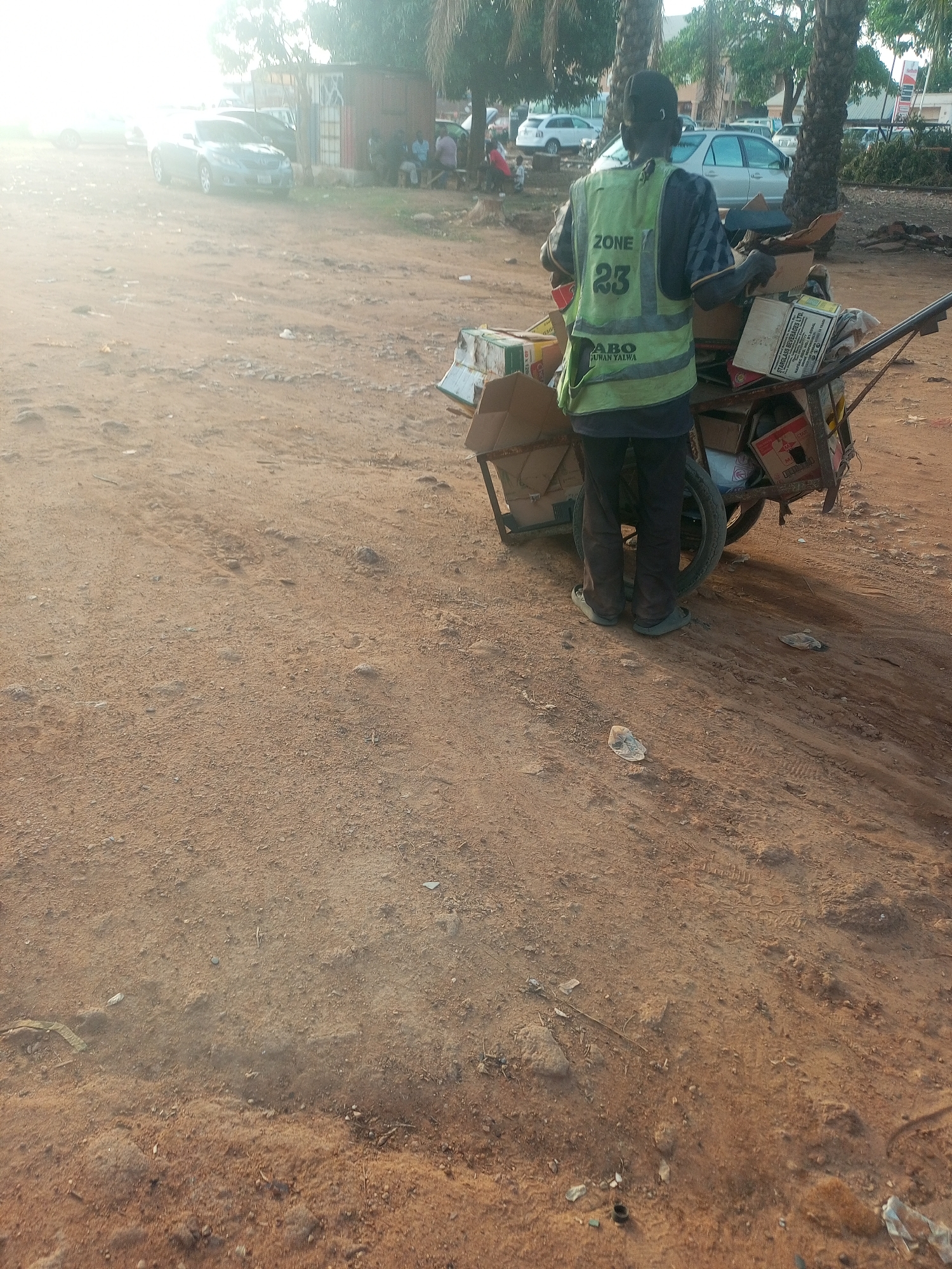 The image depicts an individual from behind, standing next to a two-wheeled metal handcart heavily loaded with assorted cardboard boxes and other items. The person wears a light green or yellow safety vest over a patterned long-sleeved shirt, dark trousers, and light-colored sandals. Visible text on the vest reads "ZONE Y 23" and "ABO DUWAN YALWA". One prominent cardboard box in the cart displays "FIBREGLASS DEVELOPERS LTD."
The scene is an unpaved, reddish-brown dirt ground, typical of an informal road or open area, with scattered small stones and some debris, including plastic wrappers, near the cart. In the background, several cars are visible, including sedans and possibly an SUV, some parked and others partially obscured by trees. Tall palm trees and other foliage are present, alongside what appears to be informal structures or buildings. A small group of people can be seen sitting under a tree or shelter further in the background.
The activity suggests the individual is engaged in collecting or organizing materials, likely for recycling or disposal, given the nature of the loaded cart. The lighting indicates either early morning or late afternoon, with bright sunlight causing a strong glare on the left side of the frame, signifying a sunny day. The overall setting, including the unpaved environment and specific text on the vest, aligns with the provided location context of Yelwa, Nigeria. No specific emotions are discernible from the back of the individual.