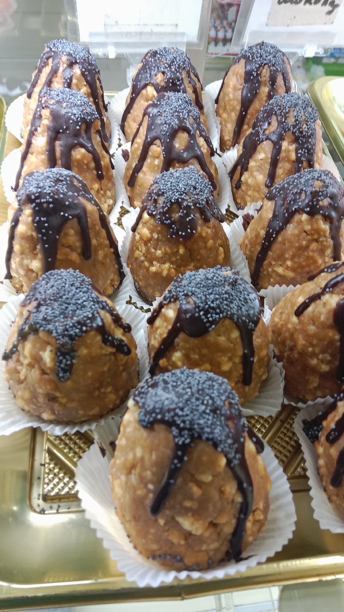 A close-up, eye-level shot shows a tray of numerous small, cone-shaped pastries with a dense, crumbly texture, possibly made from nuts and cookie crumbs. The pastries are topped with a dark chocolate glaze drizzled down the sides and sprinkled with poppy seeds. They are individually placed in white paper cupcake liners, arranged on a golden-colored tray. The background is blurred, suggesting a display case, with hints of other food items and signage. The scene appears to be a commercial setting, likely a bakery or confectionery shop in Petropavl, Kazakhstan, given the context of the location. There are no people visible in the image.