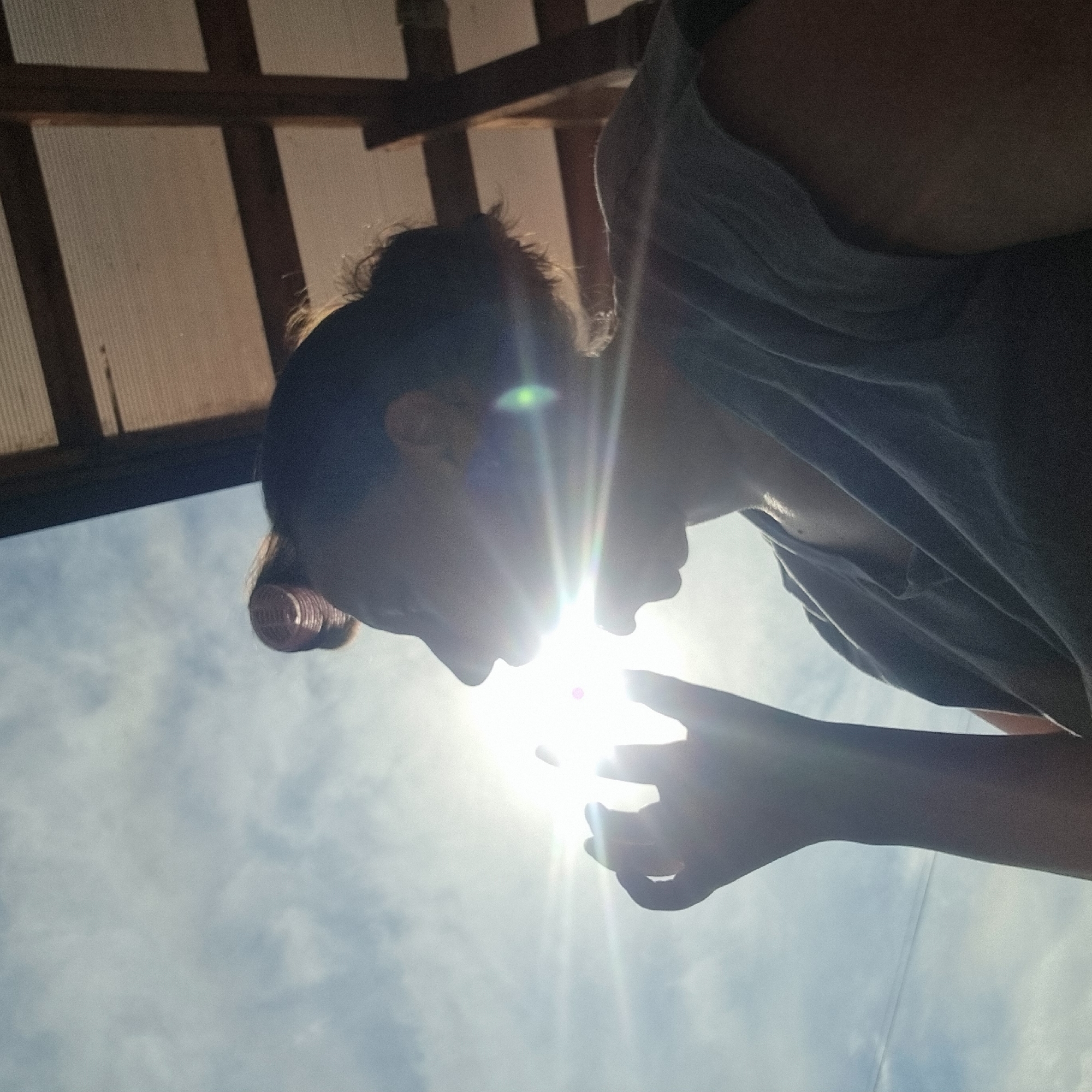 The image shows a close-up, low-angle view of a person silhouetted against a bright sky. The sun is directly in the frame, creating lens flare and overexposure, obscuring much of the detail. The person's head is tilted, with their profile visible. Their hair appears to be styled with rollers. A hand is raised, partially covering the sun. The person is wearing a dark-colored t-shirt. In the background, a structure made of wood and corrugated material is visible, suggesting an outdoor shelter or roof. The sky is partly cloudy. The location is specified as Dunnae-myeon, South Korea.