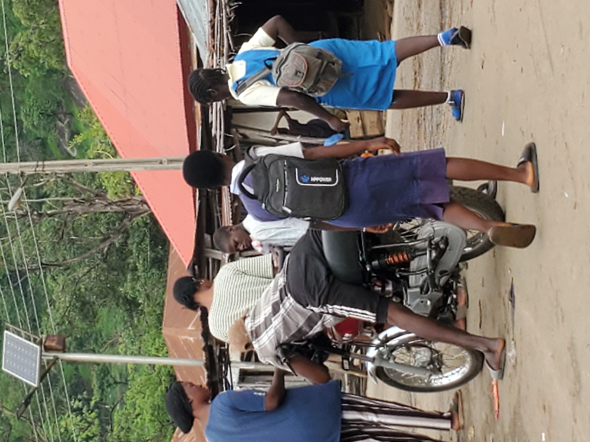 The image depicts a scene in Mararaba, Nigeria. A motorcyclist is seated on his motorcycle, with two school girls appearing to be passengers. The girls are wearing school uniforms and backpacks. Nearby, another adult is seen looking up towards the motorcycle. A solar panel is visible to the left of the scene. The location appears to be an urban area with buildings and trees.
