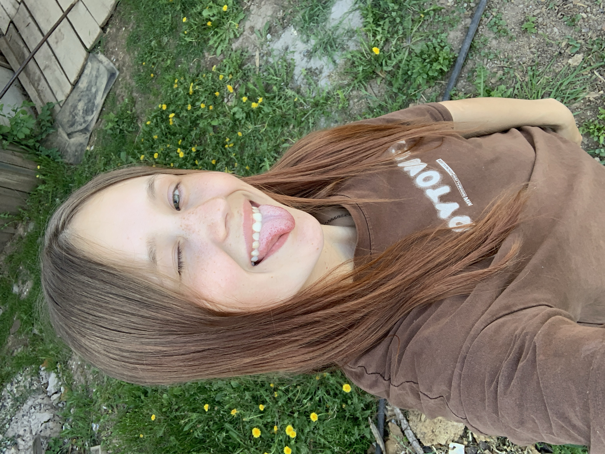 A young woman is taking a selfie. She is sticking her tongue out and winking. She has light skin, freckles, and brown hair. She is wearing a brown t-shirt with white lettering. The background is a grassy area with yellow flowers. A wooden structure is visible in the upper left corner. It is daytime. The location is unknown, but the city is Фоки, Russia.