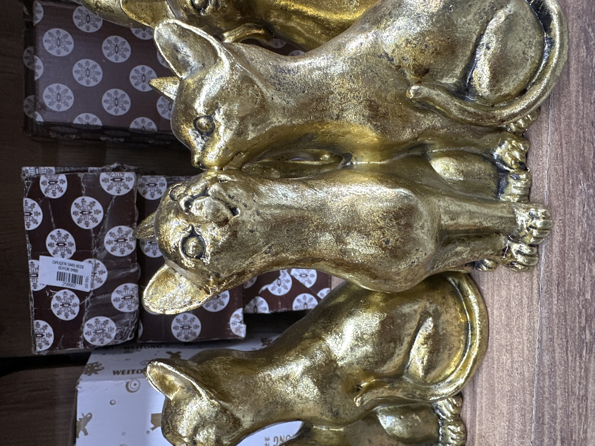 The image shows three golden cat figurines stacked on top of each other, sitting on a brown wooden surface. They appear to be resting on boxes with decorative brown and white patterns. One box also has a white label with "OPUSEN SARI KEDI BUYUK 0480" printed on it. Another box has the word "WEITONG" printed on it. The location is suggested as Fethiye, Turkey, but this is not visually verifiable from the image alone.