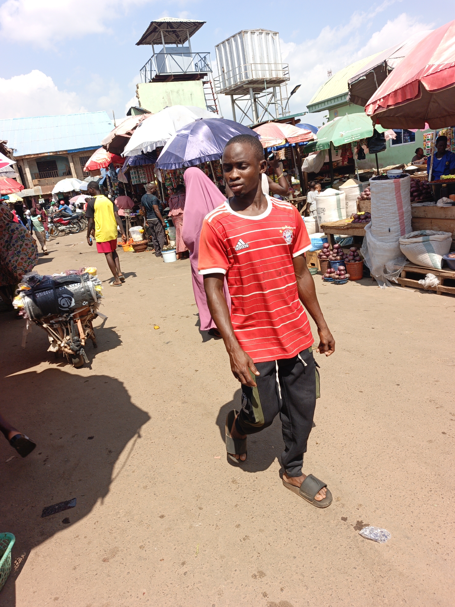 A man wearing a red and white striped Adidas shirt, black sweatpants, and sandals walks on a paved road in an outdoor market in Akwanga, Nigeria. The market is bustling with people, goods, and vendors. The ground is partially shaded. In the background are various stalls covered with umbrellas and canopies. A water tower and a raised platform structure are visible in the distance. Other individuals are present in the background, including people shopping, selling goods, and walking around. A loaded cart is on the left of the scene.