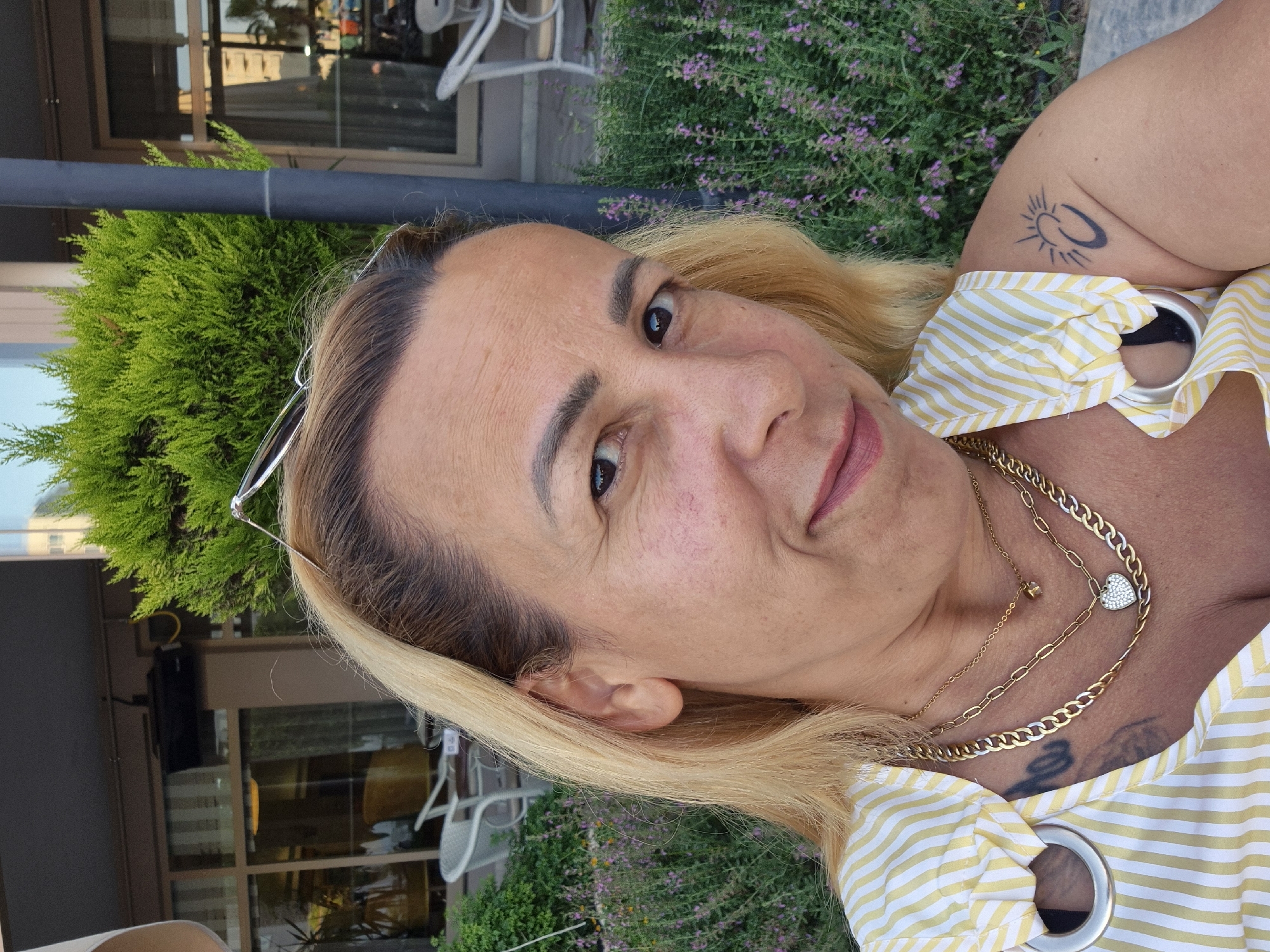 A medium shot captures a woman from the shoulders up, with her face turned slightly to the right. She has blonde hair and is wearing a yellow and white striped top with gold necklaces. The background shows a lush green bush and some buildings. The woman's left shoulder bears a tattoo of a sun and moon. The lighting suggests it is daytime. The location is identified as Unknown, Unknown.