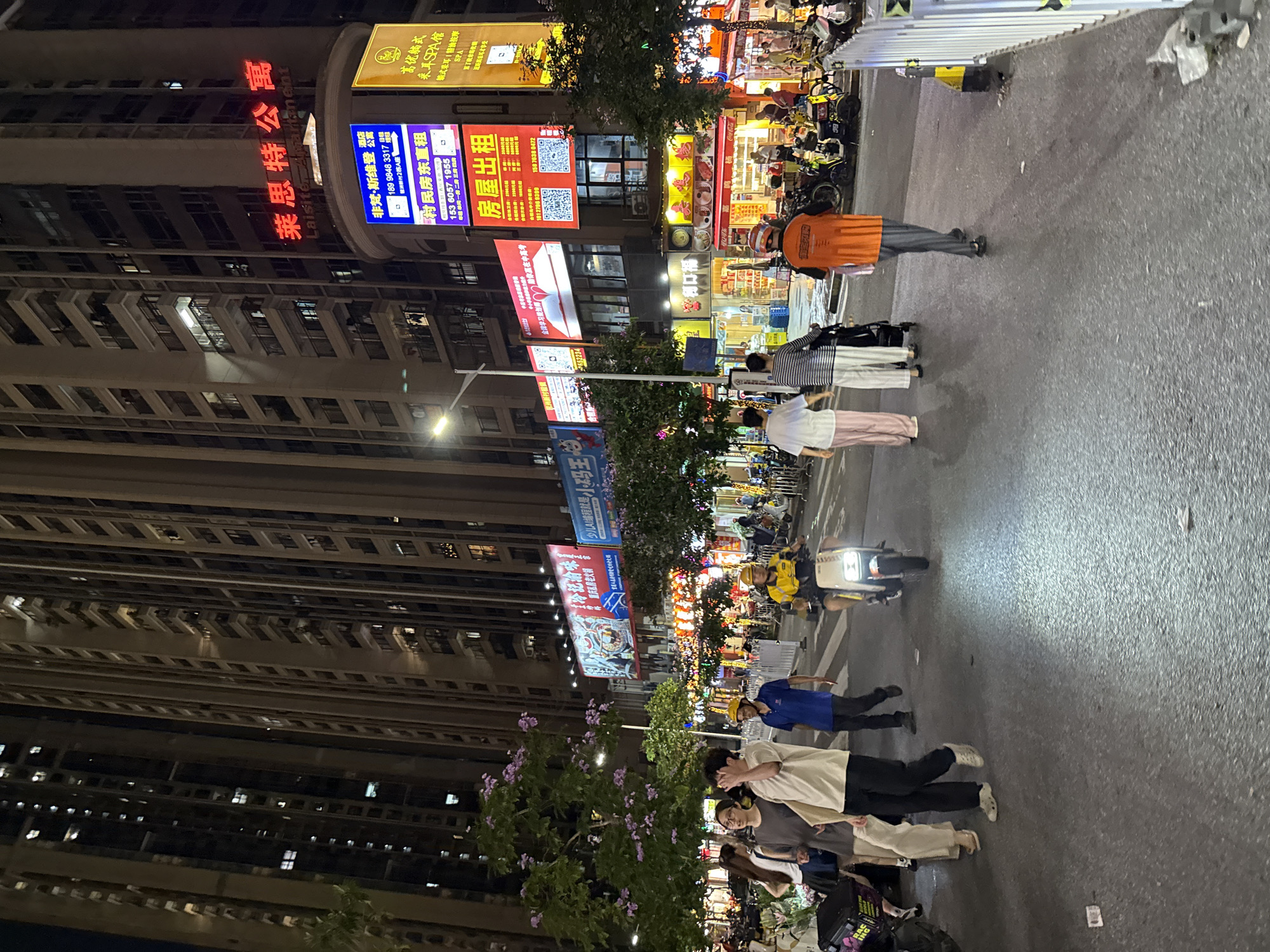 The image shows a street scene at night in Haizhu District, China. Tall apartment buildings are visible on the left side of the frame, adorned with neon signs. The street is active with pedestrians and a moped. Several storefronts with brightly lit signs and displays are along the right side of the street. There are many people visible, walking, and driving.