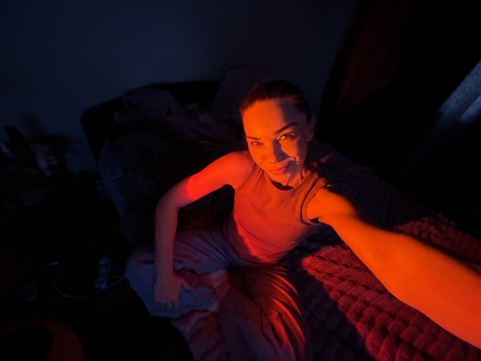 A woman with dark hair pulled back is seated on a bed covered with blankets. She is wearing a grey tank top and light-colored pants. The scene is illuminated by red and blue light, creating dramatic shadows. Her right arm is extended towards the camera, and she is looking directly at the lens with a slight smile. The background is dark and indistinct, with some crumpled fabric visible. The environment appears to be indoors, possibly a bedroom.