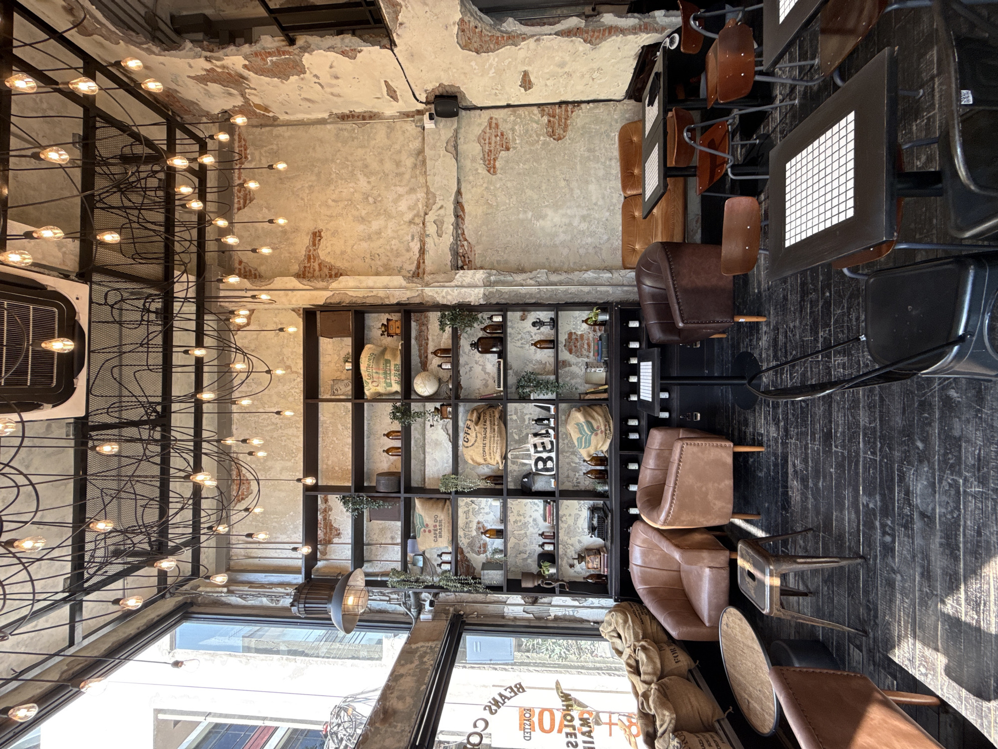 The image depicts the interior of a coffee shop in Bangkok, Thailand, characterized by an industrial-rustic aesthetic. The scene is devoid of people. The left side features large floor-to-ceiling windows, through which a mirrored sign advertising "BEANS COFFEE ROASTED WHOLESALE & RETAIL MAIN" is partially visible. Sacks of coffee beans rest on the dark wooden floor near the windows.
The walls are a distressed combination of exposed red brick and peeling light-colored plaster, revealing the underlying structure. The ceiling features exposed dark metal beams and numerous bare, suspended Edison-style light bulbs with visible wiring, creating a dense overhead lighting arrangement. A large, dark pendant light hangs near the window, and an air conditioning unit is mounted on the ceiling.
Along the back wall, a tall, dark wooden shelving unit is filled with various items, including burlap sacks labeled "CAFES DO BRASIL" and "COFFEE TRADE FACTORY," clear and amber glass bottles, small potted plants, books, a globe, a vintage typewriter, and a coffee grinder. A sign spelling "BELAA" is also on the shelf.
Seating arrangements include several brown leather armchairs (two lighter, one darker), a dark brown leather two-seater sofa, and multiple dark tables with white grid-patterned surfaces. These tables are paired with dark metal chairs, some featuring wooden backs. A small, round wooden table and a metallic stool are also present. The floor is dark, distressed wood, showing wear. A black speaker is mounted on the right wall.