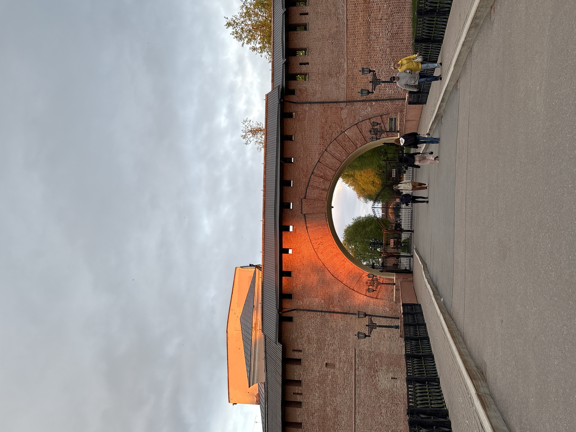 The image shows a view of the Novgorod Kremlin's walls and an archway in Veliky Novgorod, Russia. The walls are made of red brick and feature a series of small windows along the top. A metal roofline runs along the top of the walls. An orange glow appears on the walls nearest the opening and on the tall structure in front of the wall. Several people are walking on the paved path in front of the archway, some of them walking out from under the arch. There are vintage-style lampposts mounted on the walls. The sky is overcast, with a gray and cloudy appearance. Through the archway, greenery and wrought iron fencing are visible. The image appears to be taken from a slightly low angle, looking up at the walls.