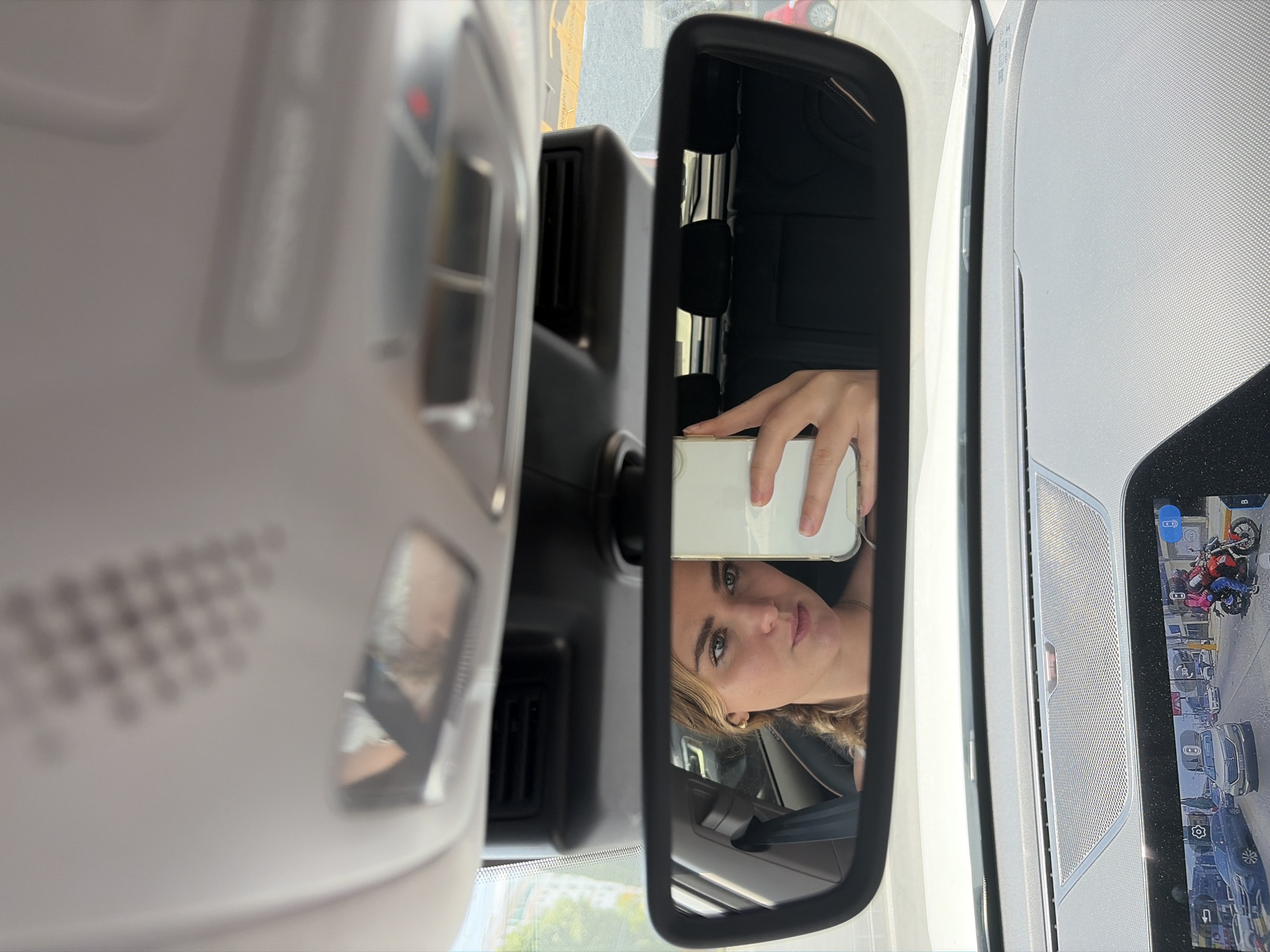 The image depicts a scene inside a car, primarily featuring a person taking a selfie in the rearview mirror. The subject, appearing to be a woman with light-colored eyes and blonde or light brown hair, is reflected from the chest up, holding a white smartphone to capture her image. Her expression is neutral or slightly pensive.
The rearview mirror is centrally positioned in the portrait-oriented frame, offering a clear reflection of the person. To the left, the car's light-colored overhead console is visible, along with air vents and blurry control elements. The vehicle appears to be modern, suggested by the sophisticated interior design and the prominent infotainment screen to the right of the rearview mirror. This screen displays a parking assist or 360-degree camera view, showing multiple vehicles—including a red and a blue motorcycle, and several cars—along with digital parking lines and small icons like "3D" and camera symbols.
Through the car's windshield and side windows, an urban or suburban environment is visible, with hints of trees and buildings under bright daylight, suggesting the photo was taken during the day. A red car is partially visible outside the vehicle on the right. The overall setting is the interior of a car, likely stationary in Puebla, Mexico, with the main activity being a personal photograph taken via reflection. No significant text is legible apart from the small icons on the infotainment screen.