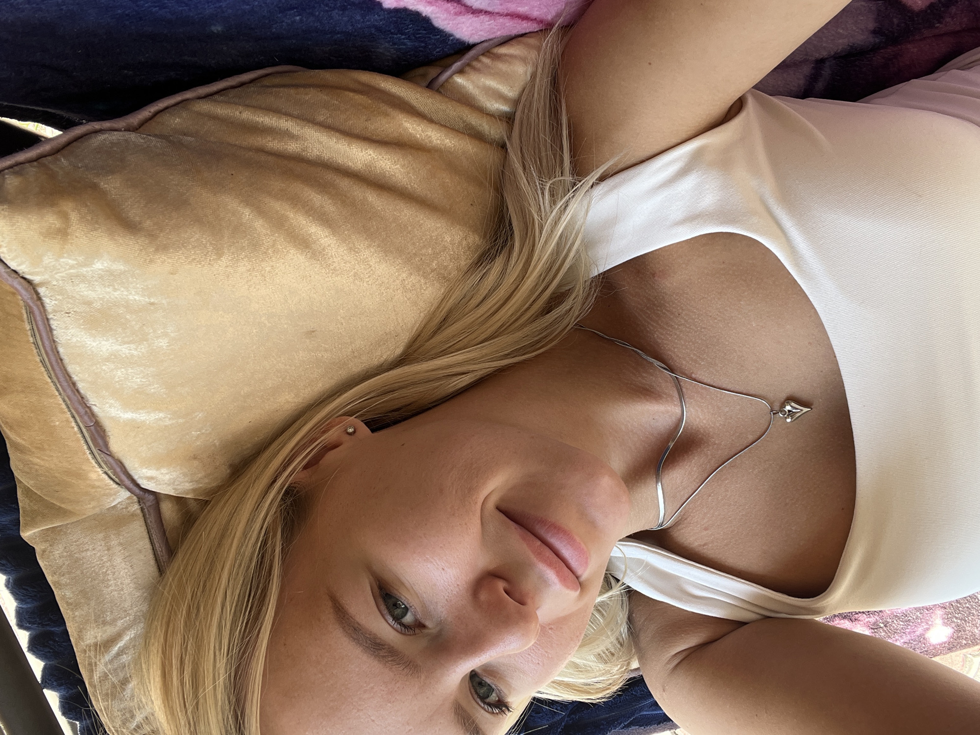 A blonde woman with fair skin and green eyes is lying down, looking up at the camera. She is wearing a white tank top and a silver necklace with a heart-shaped pendant. Her hair is blonde and styled down, partially obscuring her right shoulder. She is lying on a tan-colored pillow and appears to be indoors, possibly in a bedroom or living room, given the presence of a pillow and a blanket in the background. The lighting suggests it is daytime.