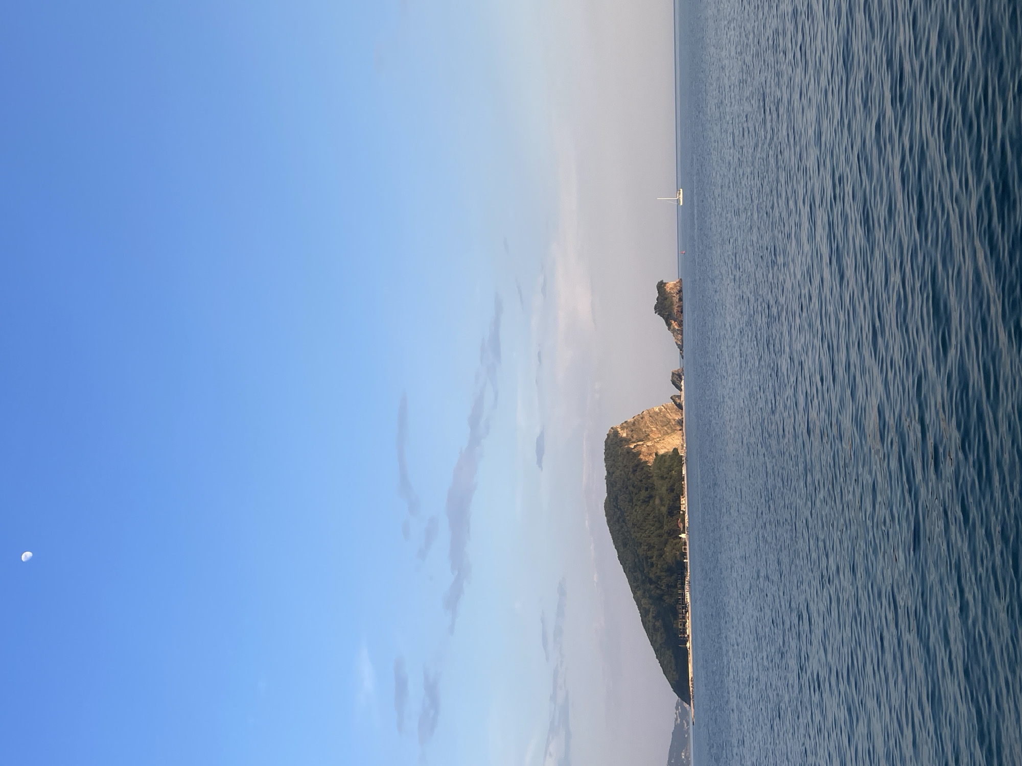 The image depicts a coastal scene, likely taken at an unspecified location. The sky transitions from clear blue on the left to a hazy, light blue-grey towards the center. A crescent moon is visible in the upper left. The sea occupies the right half of the image, its surface textured by gentle waves. An island covered in dense vegetation, with exposed rock formations, stretches from the lower center towards the top. A small sailboat is visible in the distance.
