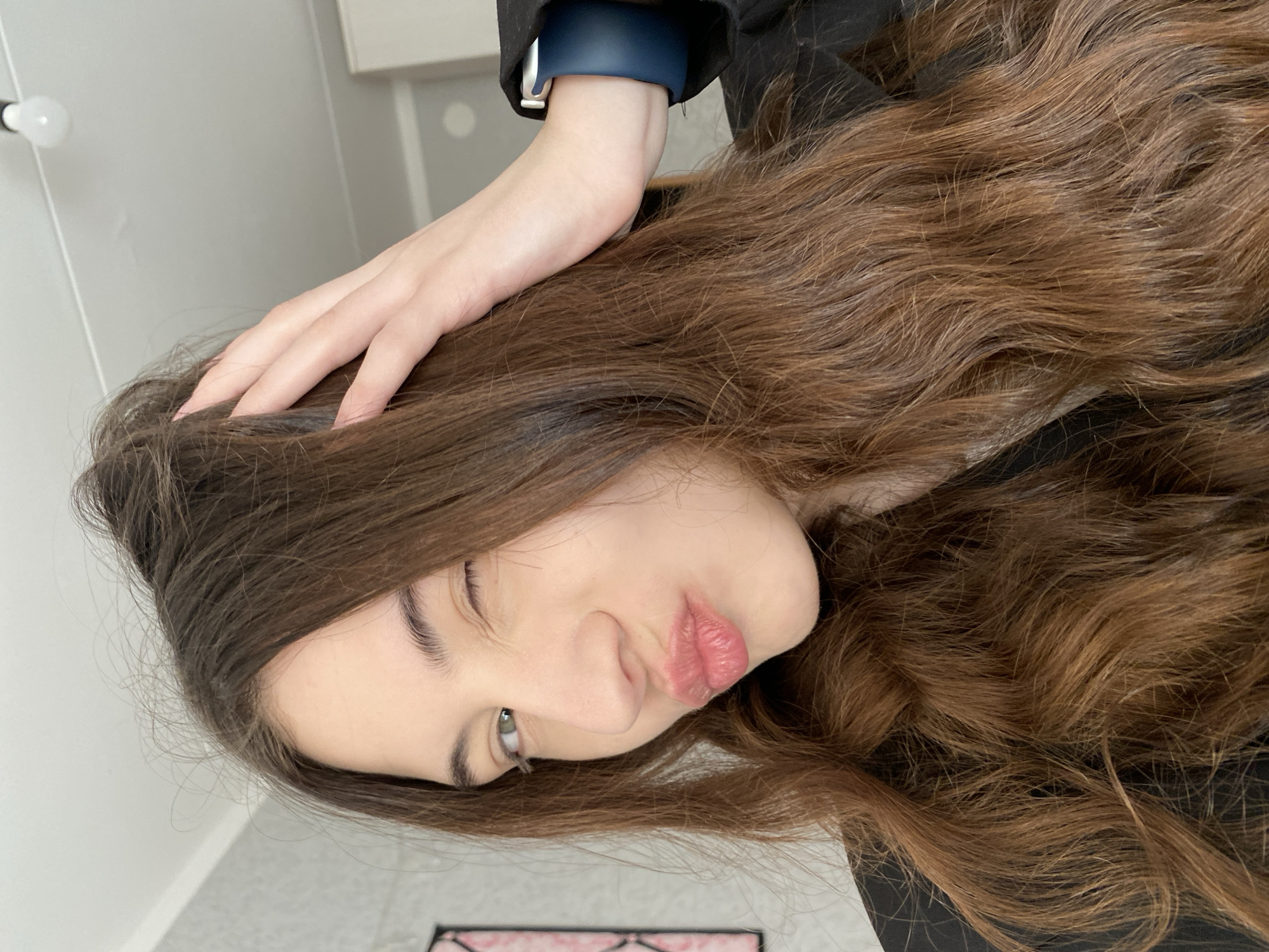 A rotated selfie of a young woman with long, wavy brown hair, wearing a dark-colored jacket and a blue smartwatch. Her head is tilted, and she is winking while pouting her lips. Her left hand rests on her head. The background includes a light bulb, a white wall with a cabinet and a tiled floor. No evidence suggests the location is in Votkinsk, Russia.
