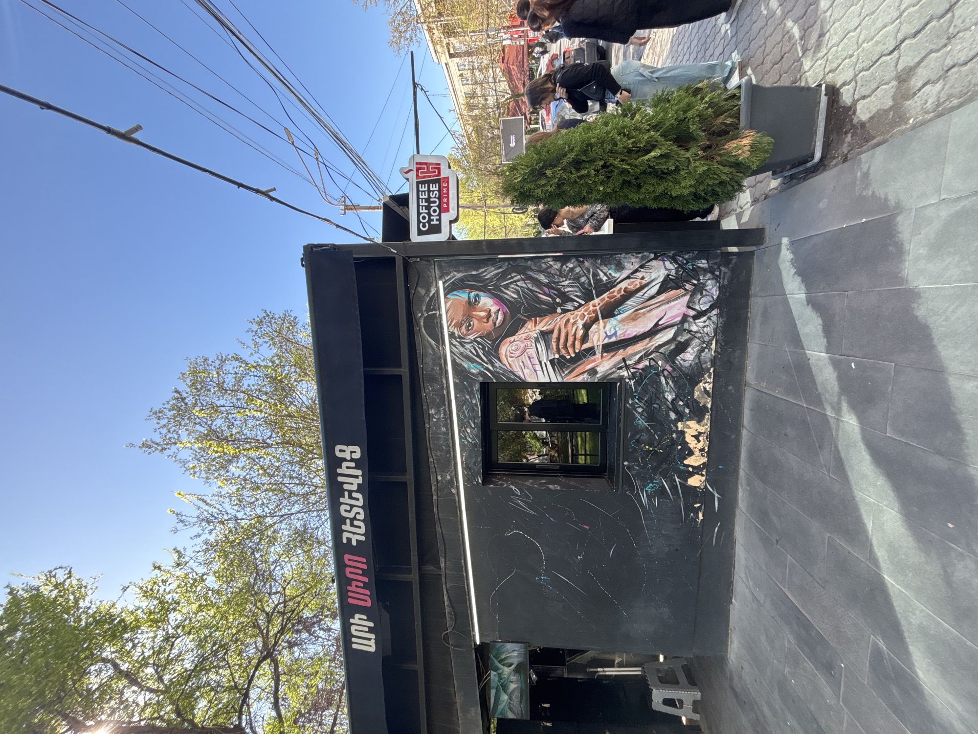 A small black building with a sign that reads "Coffee House Prime" in Yerevan, Armenia, is covered in graffiti of a woman with tattoos. Two small green trees sit in front of the building. A group of people stand on the sidewalk in the background. The sky is blue and sunny.