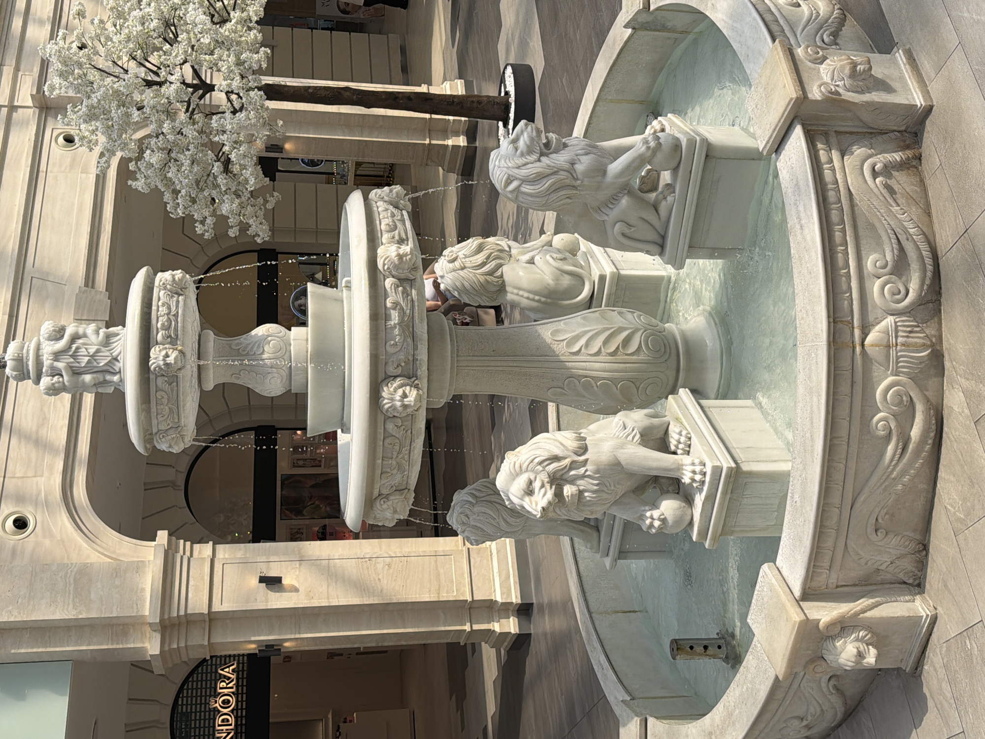 The image depicts a decorative marble fountain with sculpted lions, cherubs, and ornate carvings, located in the Atrium of the TsUM Department Store in Kyiv, Ukraine. The fountain is situated in an open-air, well-lit area with a tiled floor and classical-style architecture, including large columns and arches, visible in the background. The water from the fountain is flowing, creating a serene atmosphere. A decorative artificial tree with white blossoms stands to the right of the fountain. In the background, store entrances and signs, including one for "PANDORA", are visible. No people are prominently featured, though a person can be faintly seen sitting in the background. The overall scene suggests a public space within a commercial establishment, designed for aesthetic appeal and relaxation.