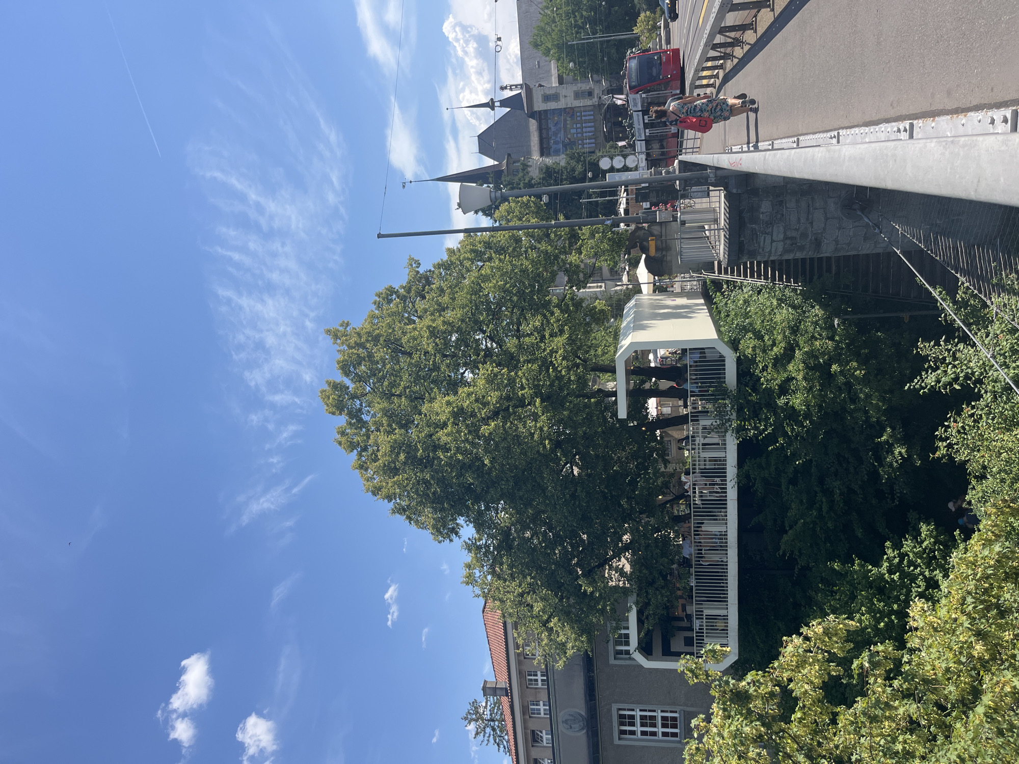 This image captures a sunny day in Bern, Switzerland, likely near the Zytglogge clock tower. In the foreground, a pedestrian bridge stretches across the frame, with a woman in a patterned dress walking away from the camera, carrying a red bag. To the left, a modern, white-paneled structure extends from a building, creating a suspended patio or seating area, populated with several people. This structure is surrounded by lush green trees. Across the street, a red tram passes by, and a building with distinctive pointed turrets, resembling a castle or historic landmark, is visible in the background. The sky is bright blue with scattered wispy clouds and a contrail from an aircraft. The scene depicts a vibrant urban environment with elements of nature and public transportation.