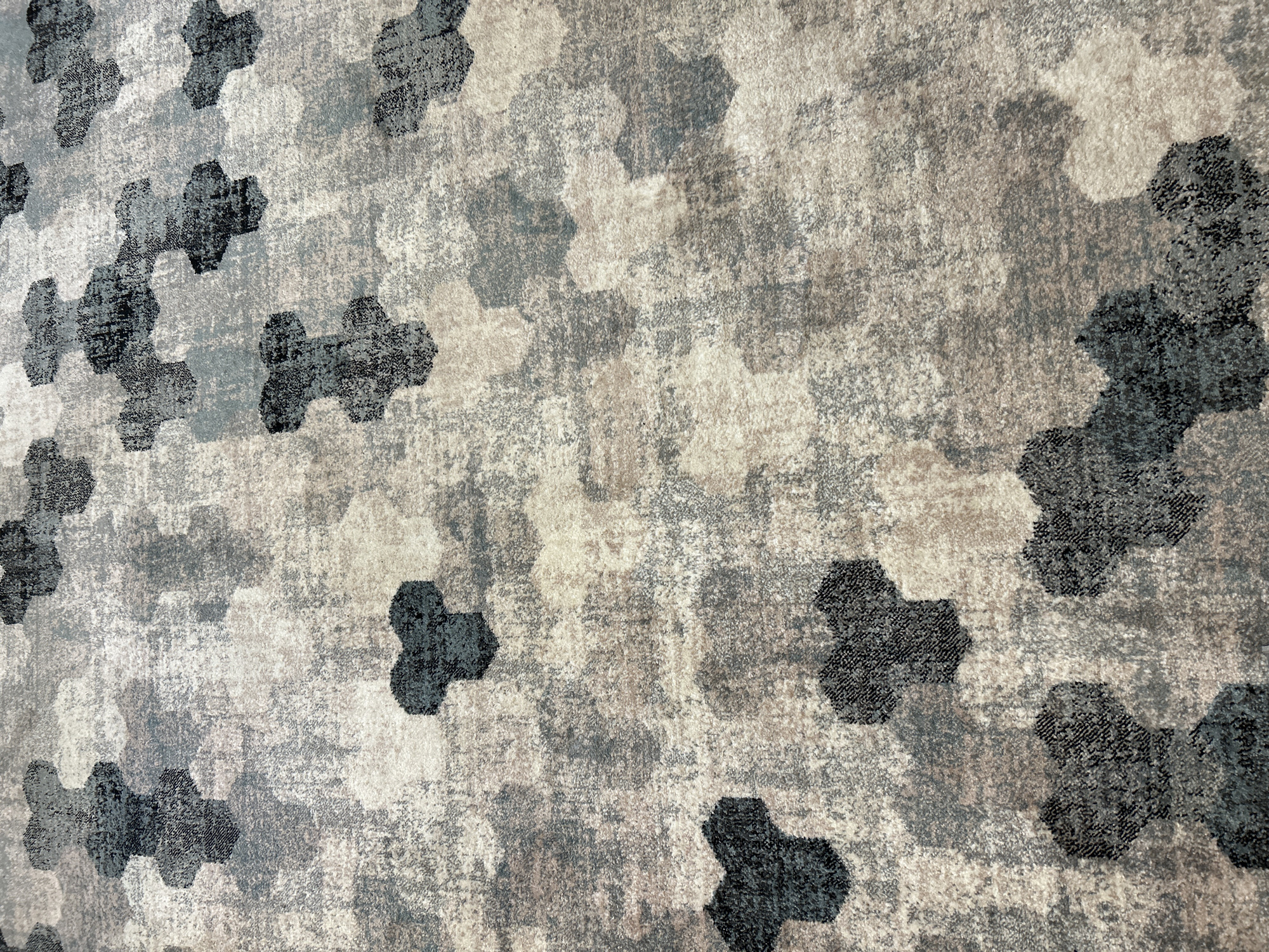 The image shows a close-up, elevated view of a textured carpet with a repeating hexagonal pattern in shades of grey, black, and beige. The hexagons appear as if they are layered or overlapping, creating a sense of depth and visual interest. The texture of the carpet is slightly rough, with individual fibers visible, suggesting it is a woven material. The lighting casts subtle shadows, highlighting the raised areas of the pattern and adding to the overall depth. There are no people, animals, or any recognizable objects other than the carpet itself. The scene appears to be indoors, likely a floor covering in a domestic or commercial space. There is no specific identifiable location within the city of Pavlohrad, Ukraine, visible in this image.