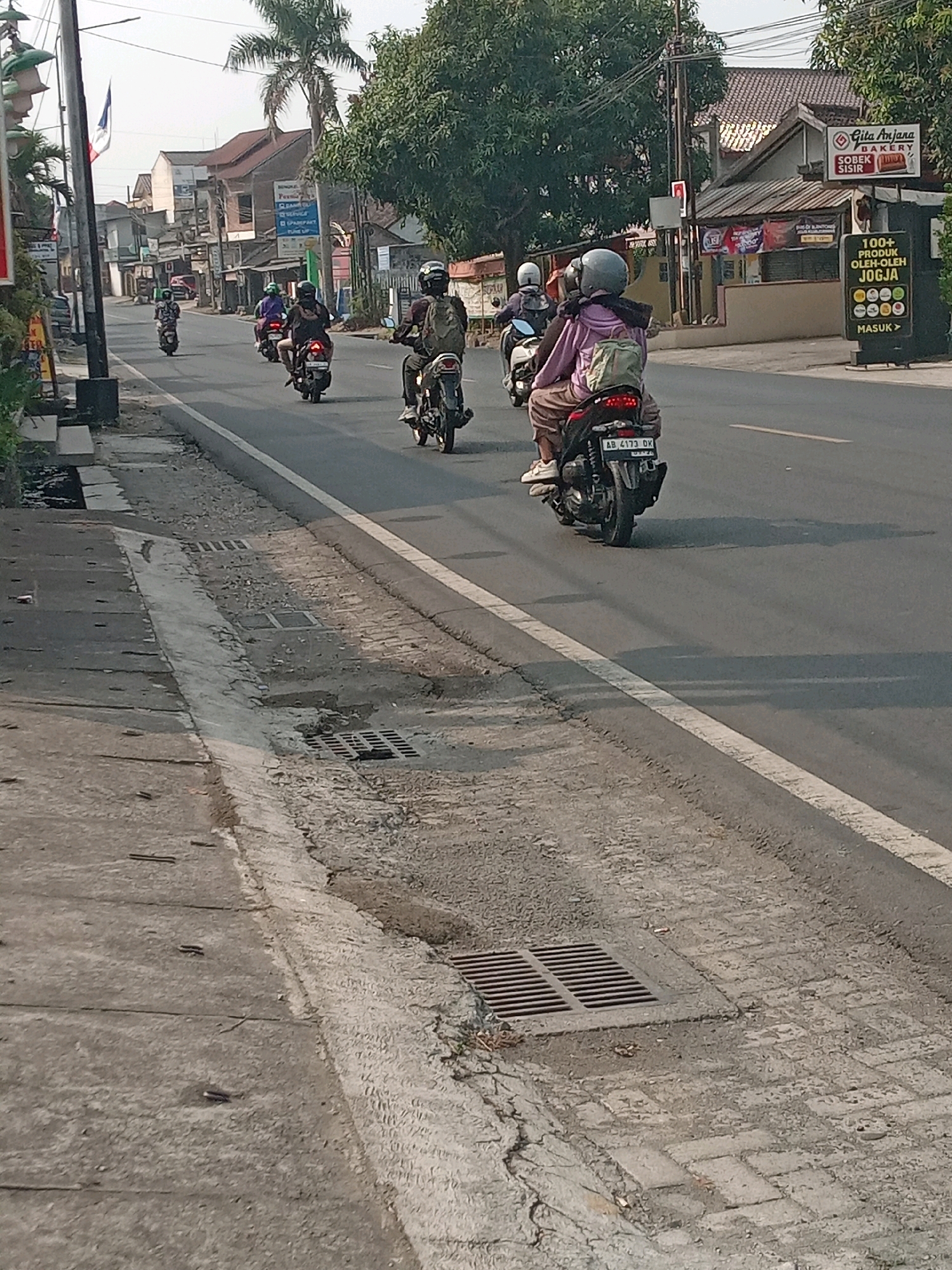 The image depicts a street scene in Banguntapan, Indonesia, with multiple motorcycles traveling on the asphalt road. In the foreground, the right side of the image shows a section of paved sidewalk and a drainage grate. The road is lined with buildings and trees. Several people are riding motorcycles, most of whom are wearing helmets. One motorcycle in the mid-ground has a license plate visible that reads "AB 4173 DK". A sign is visible on the right side of the road advertising "GITA ANJANA BAKERY" and "100+ PRODUK OLEH-OLEH JOGJA". The sky is overcast, and the lighting suggests it is daytime.