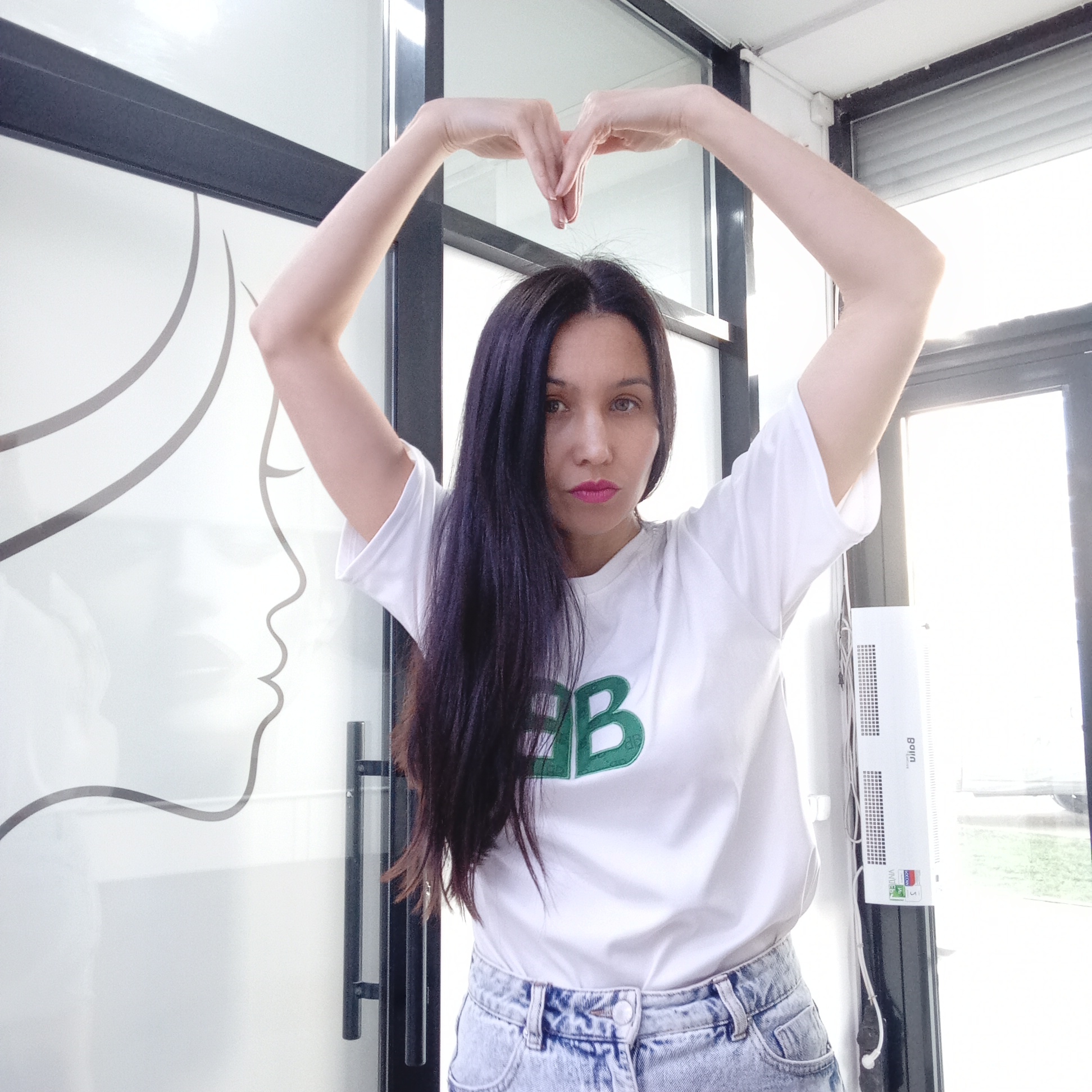 A woman is standing indoors. She is wearing a white t-shirt with a green logo and light blue jeans. She has long, dark hair. She is making a heart shape with her hands above her head. Behind her, there is a door with a frosted glass panel featuring a stylized outline of a woman's face. The room appears to be a beauty salon or clinic, judging by the logo and design on the glass. A white air conditioner is mounted on the wall next to a door. Outside the door, there is a glimpse of greenery and the roof of a car. The text “Izhevsk, Russia” has been provided as context for location.